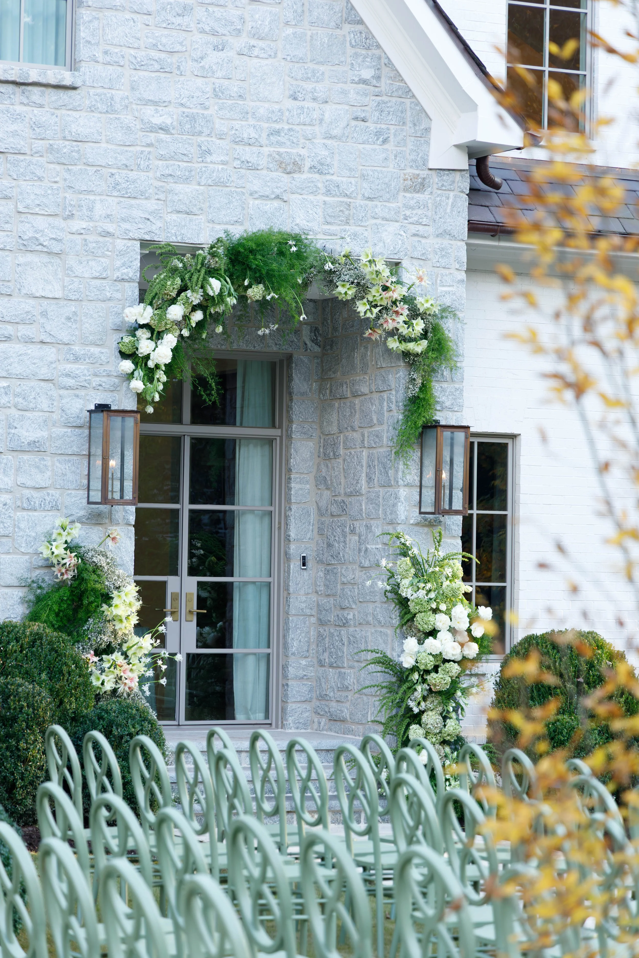 Front entrance of a house decorated with white flowers and greenery for a wedding, with rows of green chairs set up outside.