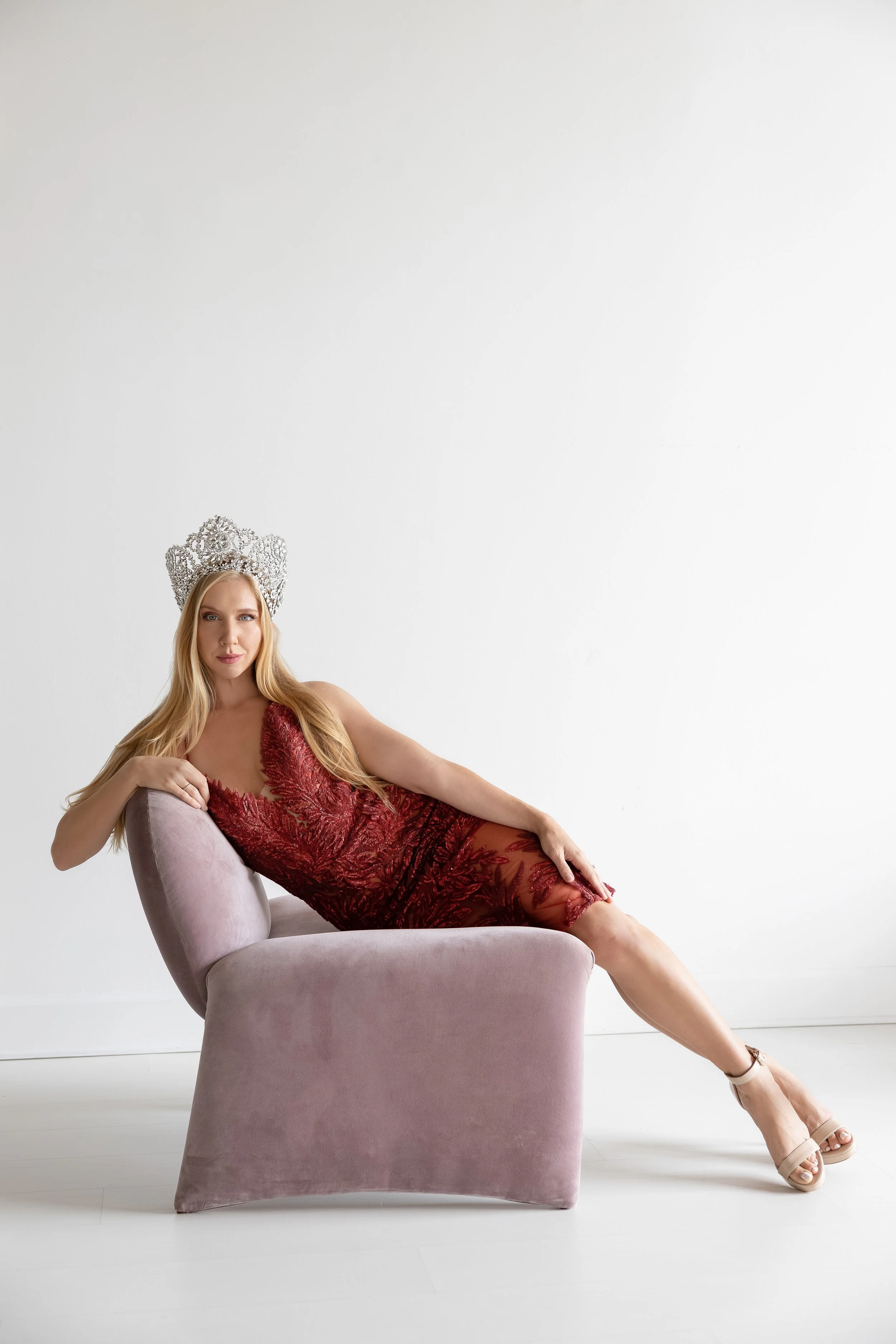 A woman lying on a pink velvet chair, wearing a red lace dress and a silver crown, with a plain white background.