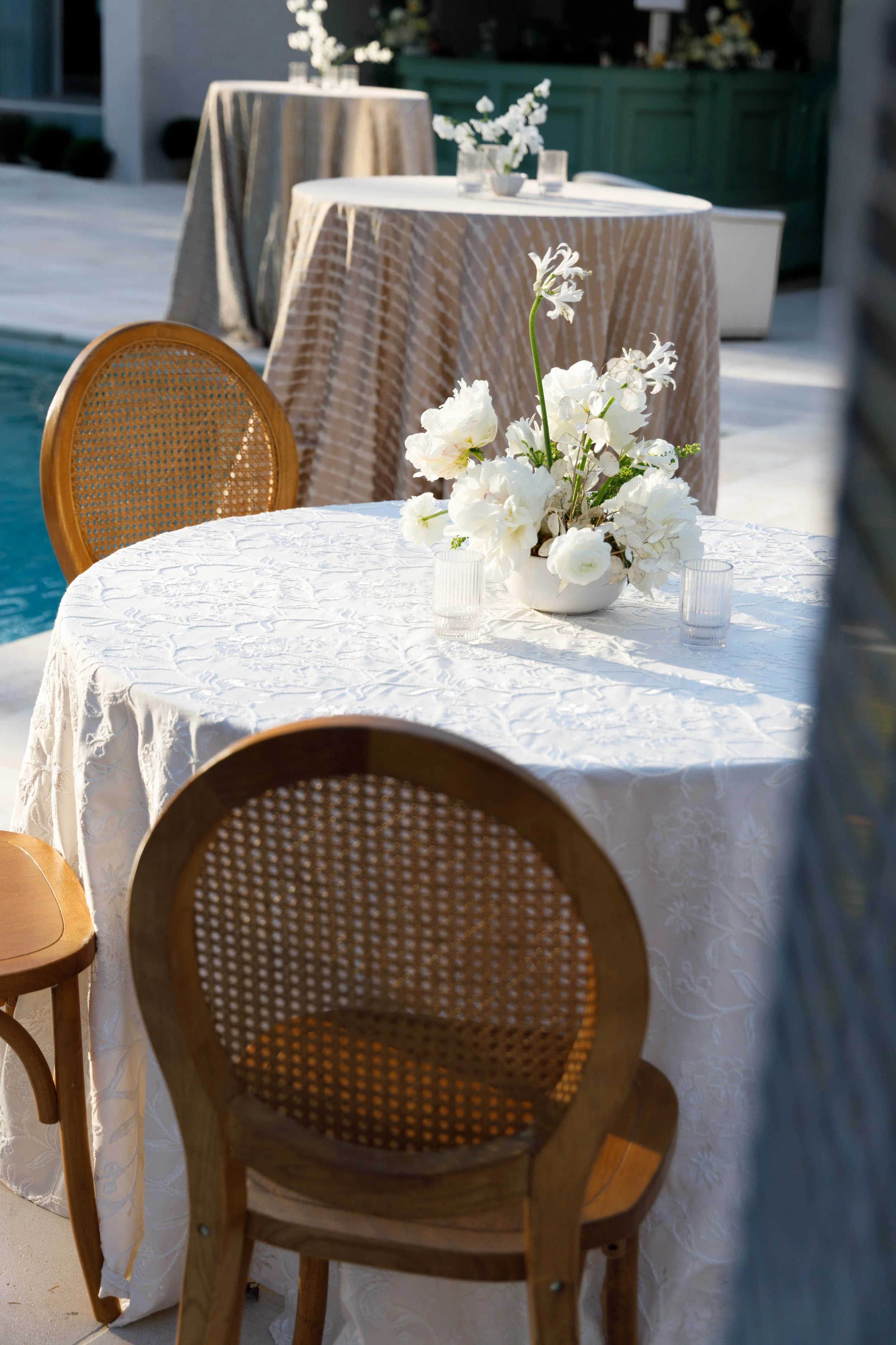 Outdoor dining table with white tablecloth, floral centerpiece, and clear glass cups, surrounded by wooden chairs with cane backs, near a pool.