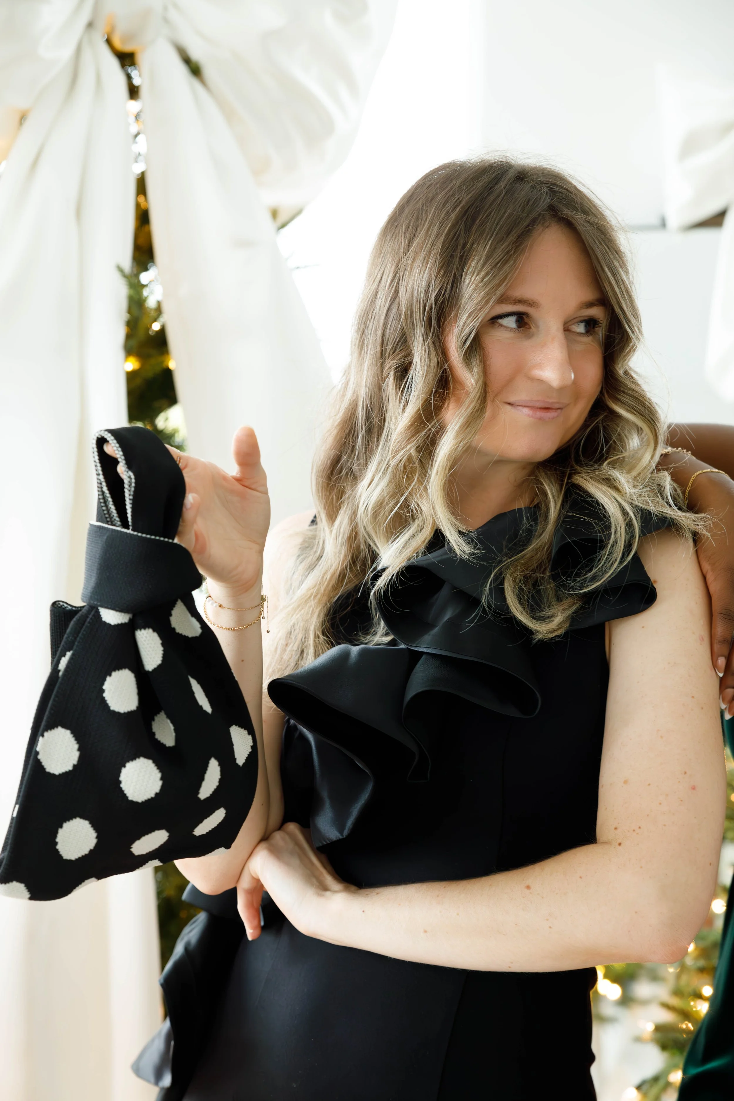 A woman with wavy light brown hair wearing a black dress with ruffled details, holding a polka dot bag, standing in front of a decorated Christmas tree and white curtains.