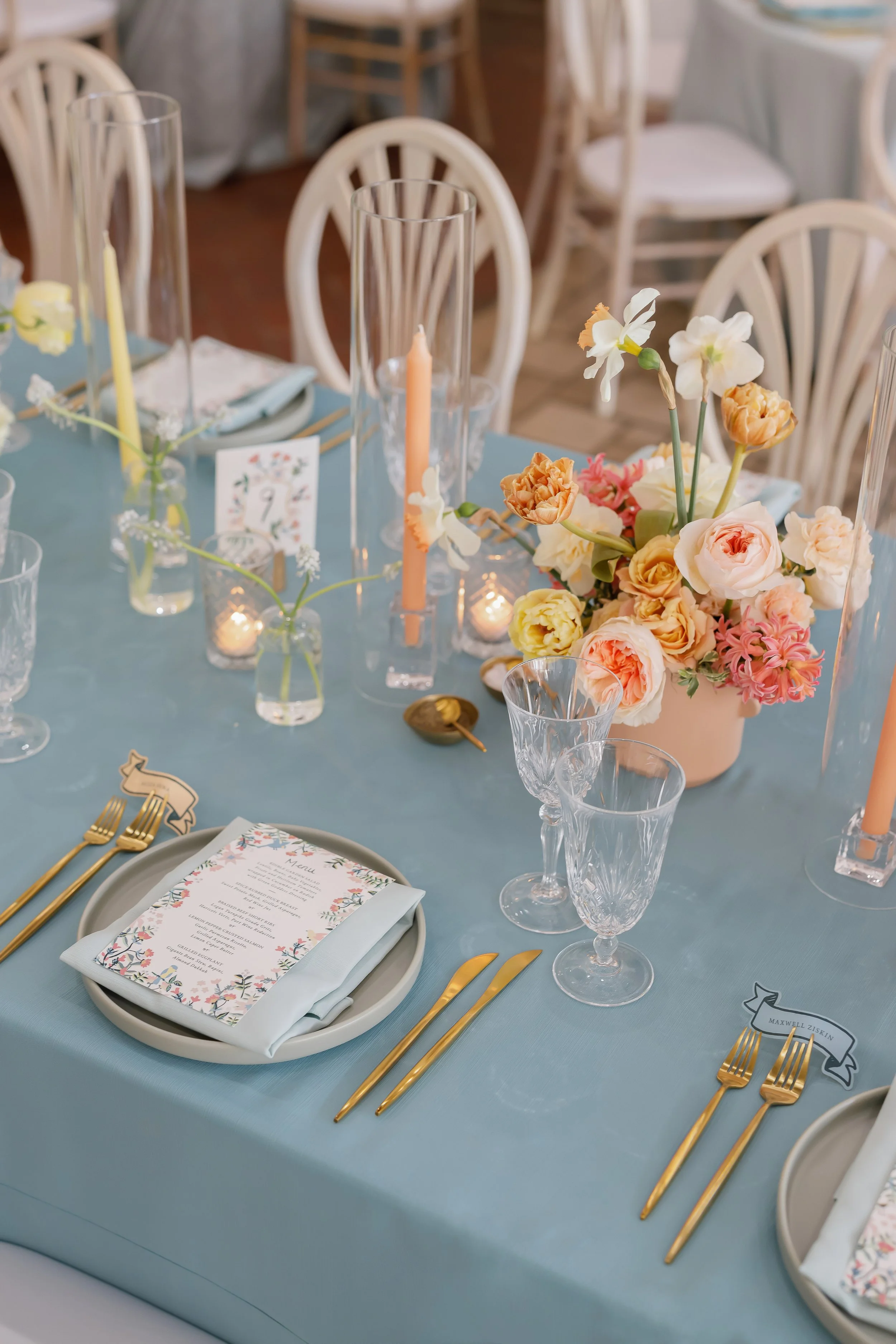Elegant dining table decorated with a centerpiece of mixed pastel-colored flowers, surrounded by glassware, tall candles, gold flatware, and printed menus on light blue tablecloths, set for a formal event.