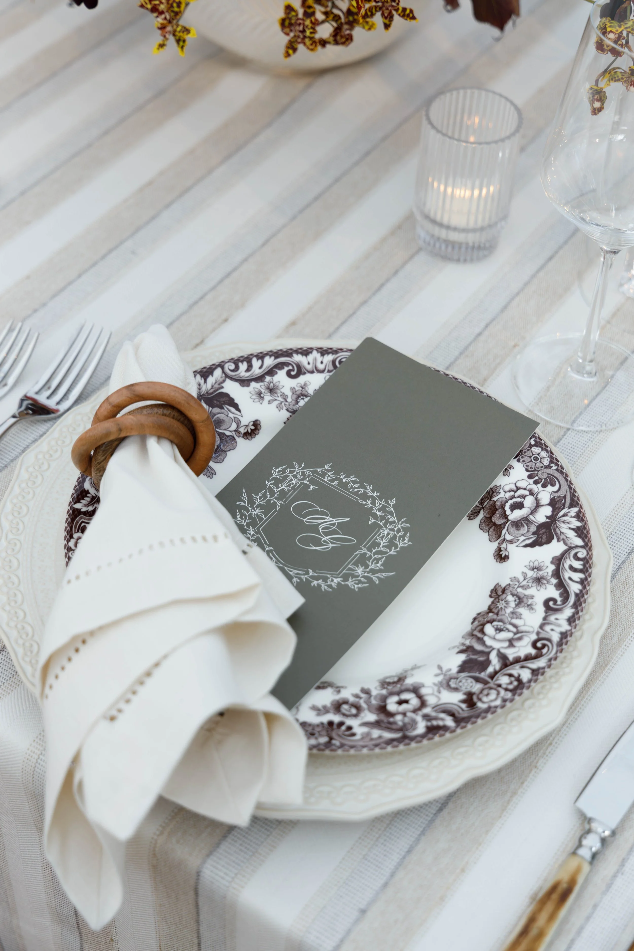 Elegant table setting with floral plate, white napkin held with wooden rings, a gray menu card, wine glass, candle, and striped tablecloth.