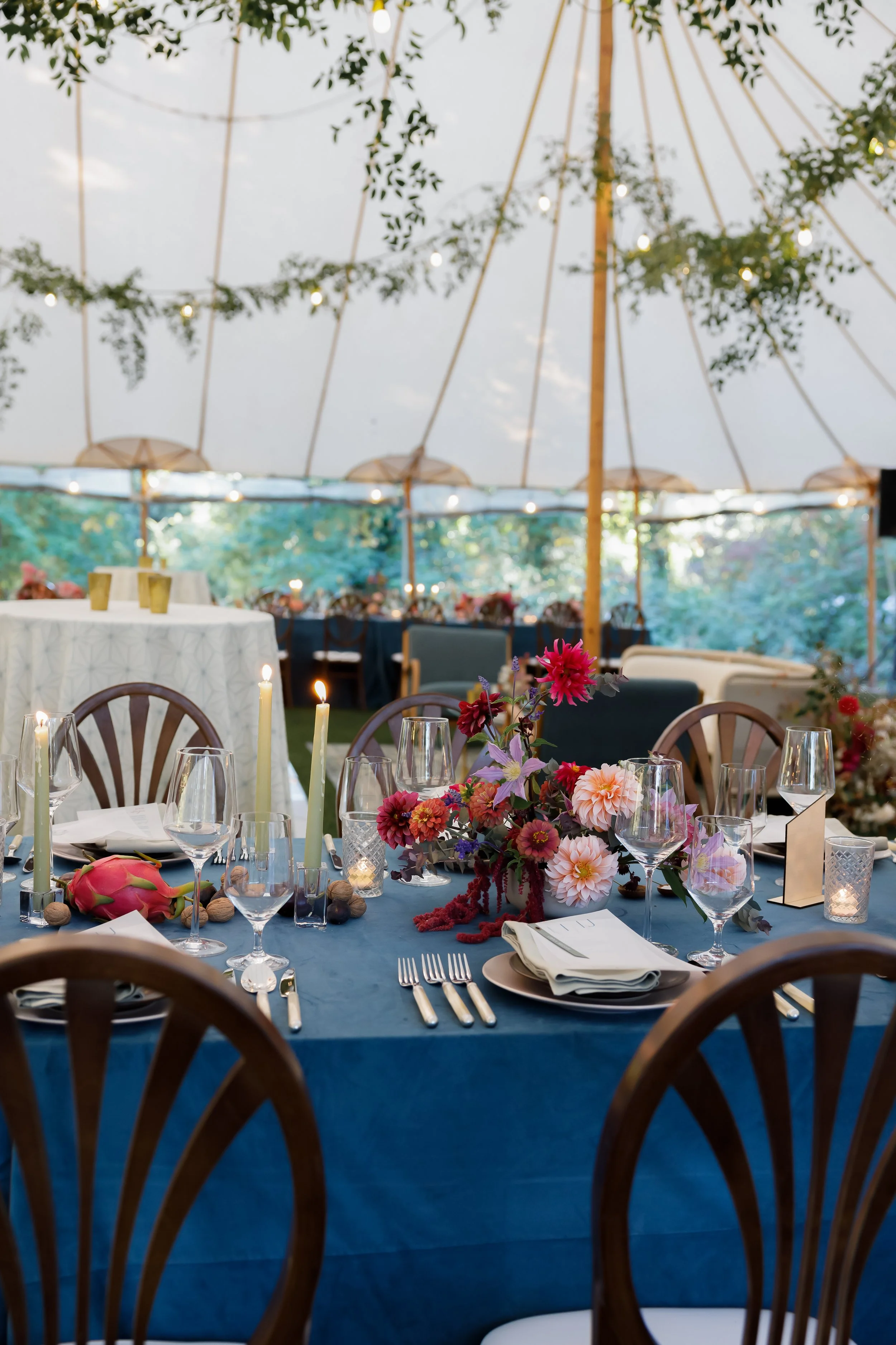 Elegant outdoor dining setup with a blue tablecloth, floral centerpiece, candles, and glassware, under a large canopy decorated with greenery and string lights.