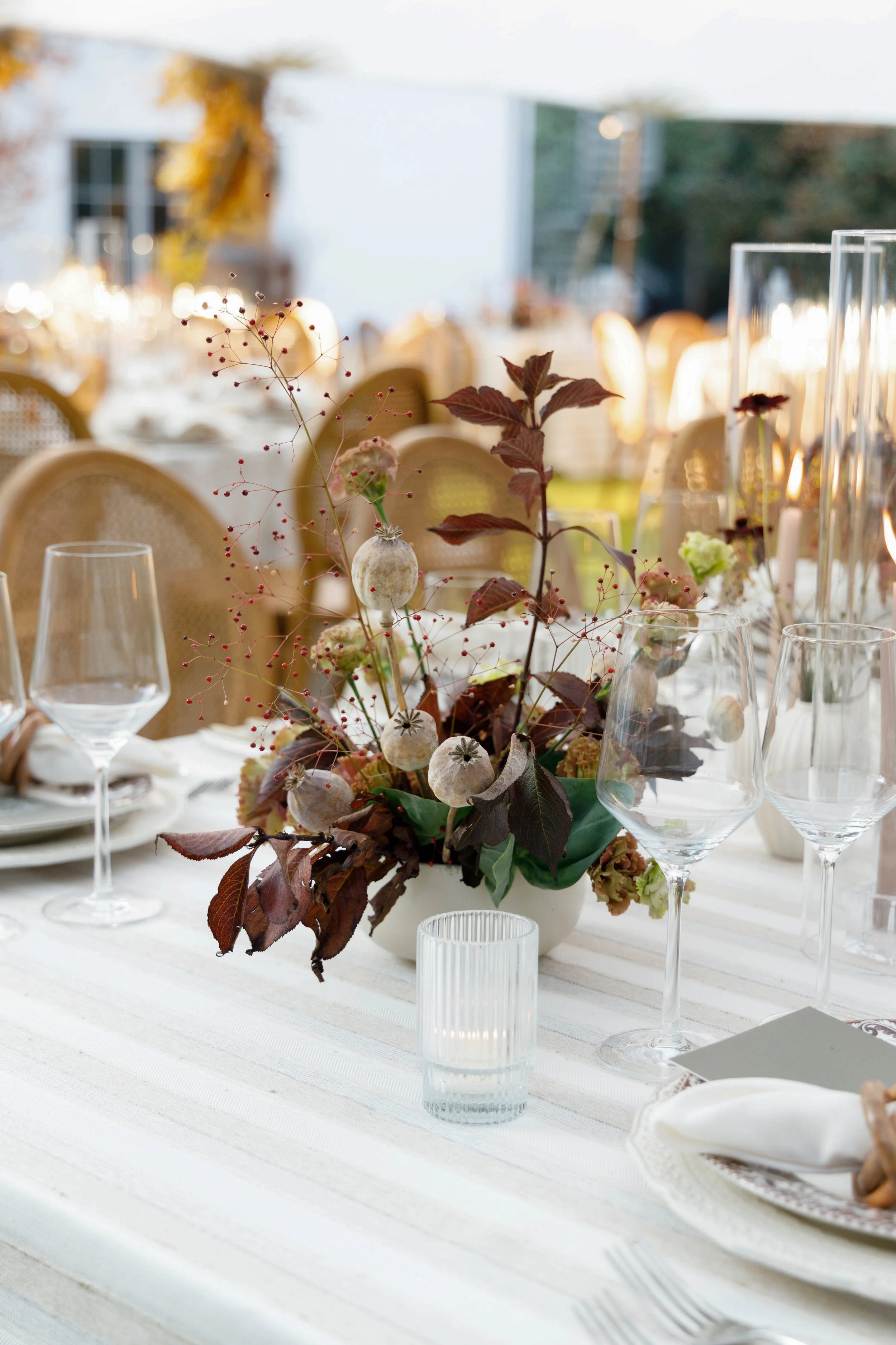 Elegant outdoor dining table decorated with a centerpiece of dried flowers and greenery, wine glasses, and candles in clear holders, set in the late afternoon sunlight.