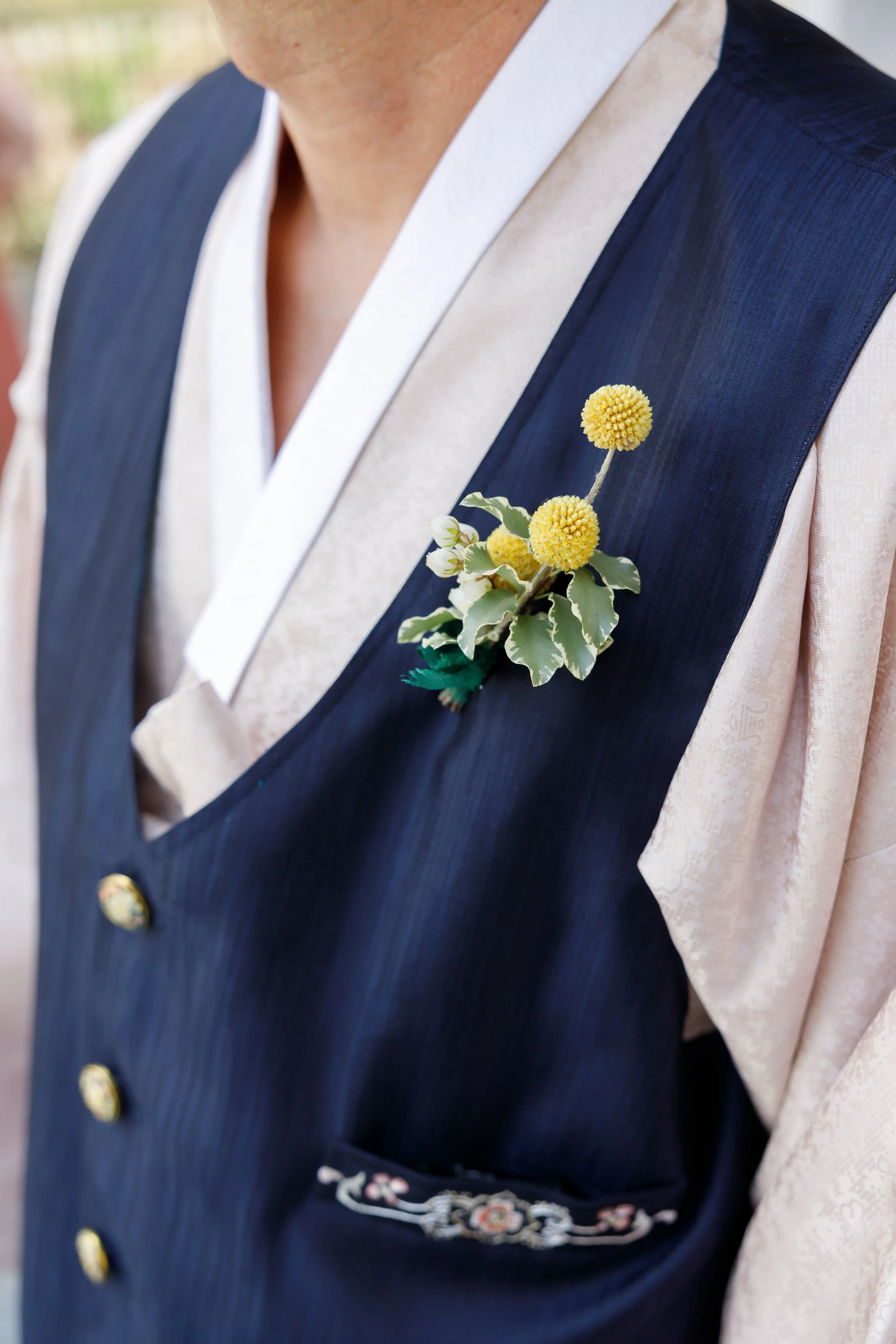 A close-up of a man's chest showing a boutonniere with yellow flowers and green leaves attached to a navy vest over a light-colored shirt.