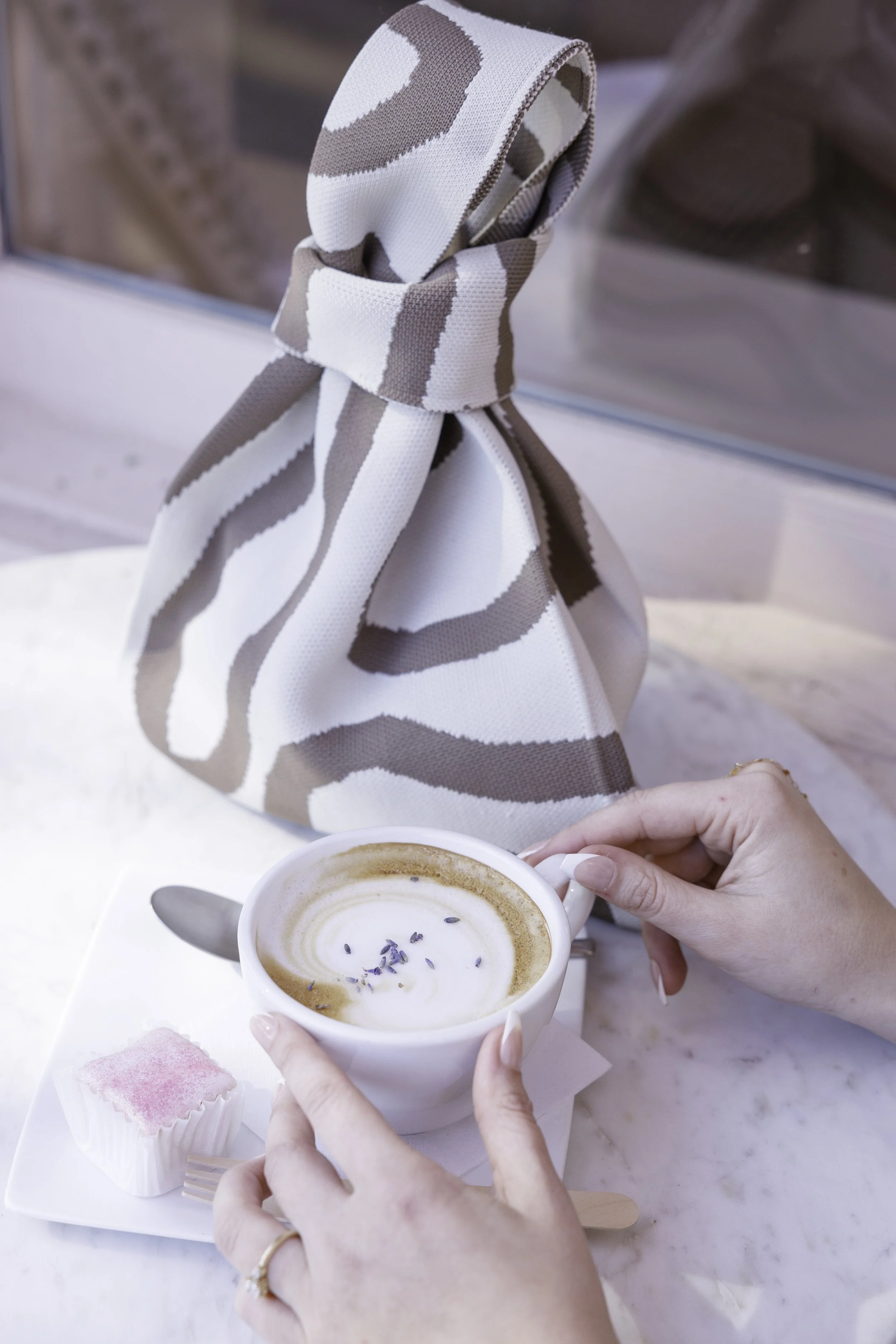 A person holding a white mug of coffee with lavender sprinkles on top, sitting at a table with a small pink dessert and a patterned bag in the background.