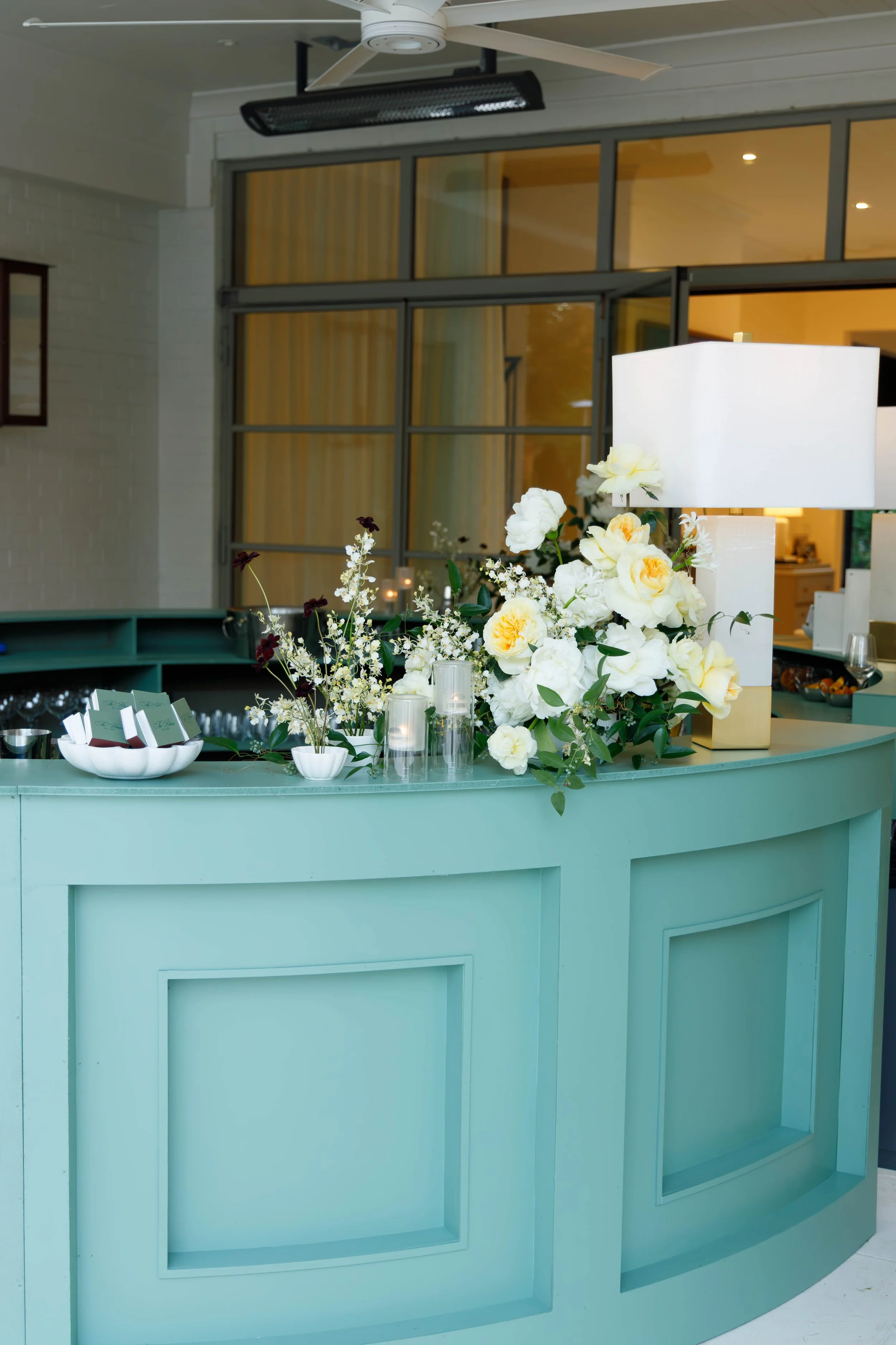 A mint green reception or bar counter decorated with white and cream flowers, a white lampshade, candles, and small bowls, in an indoor setting with large windows and ceiling fan.