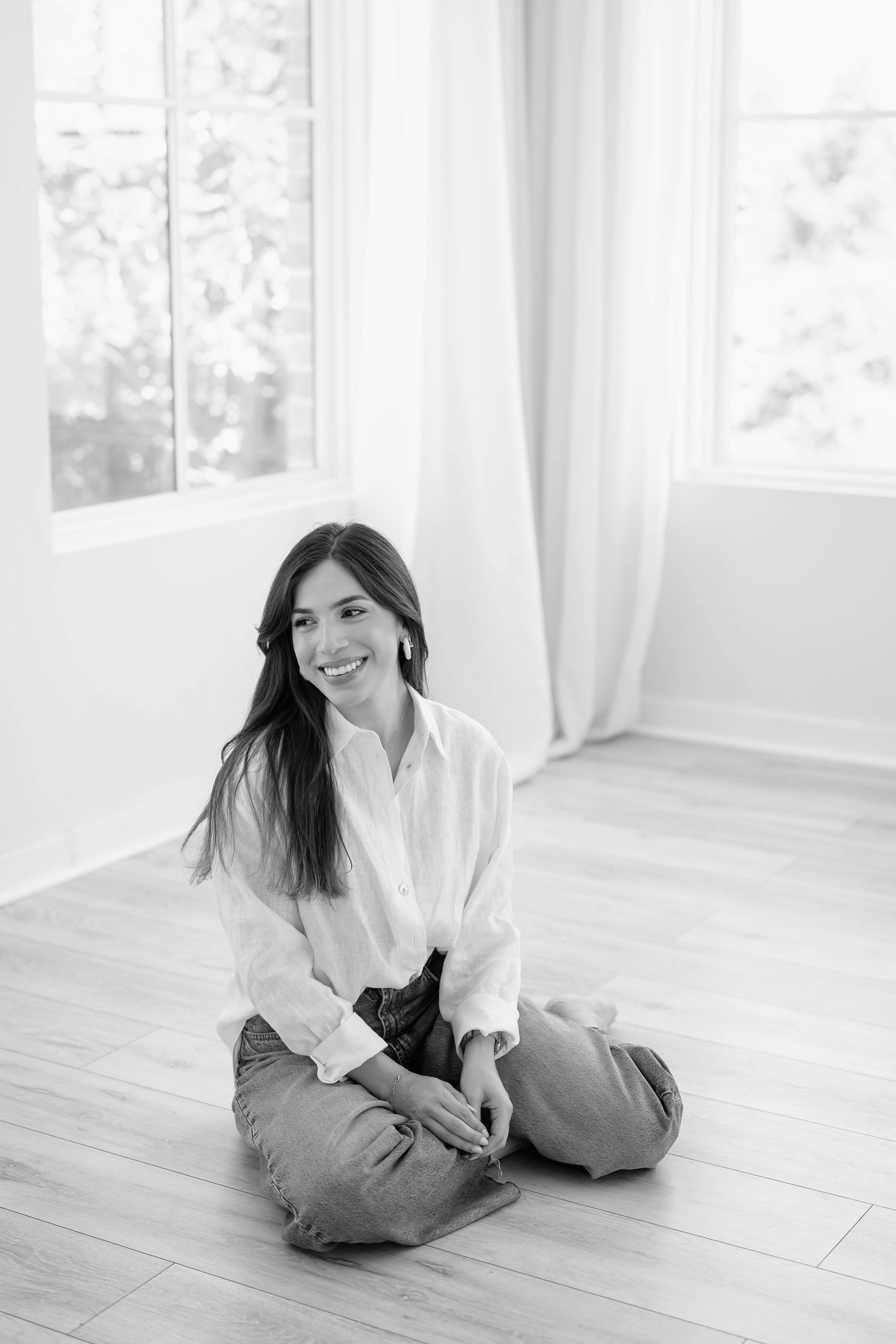 A woman sitting cross-legged on a wooden floor in a bright room with large windows and white curtains, smiling and looking to her left.