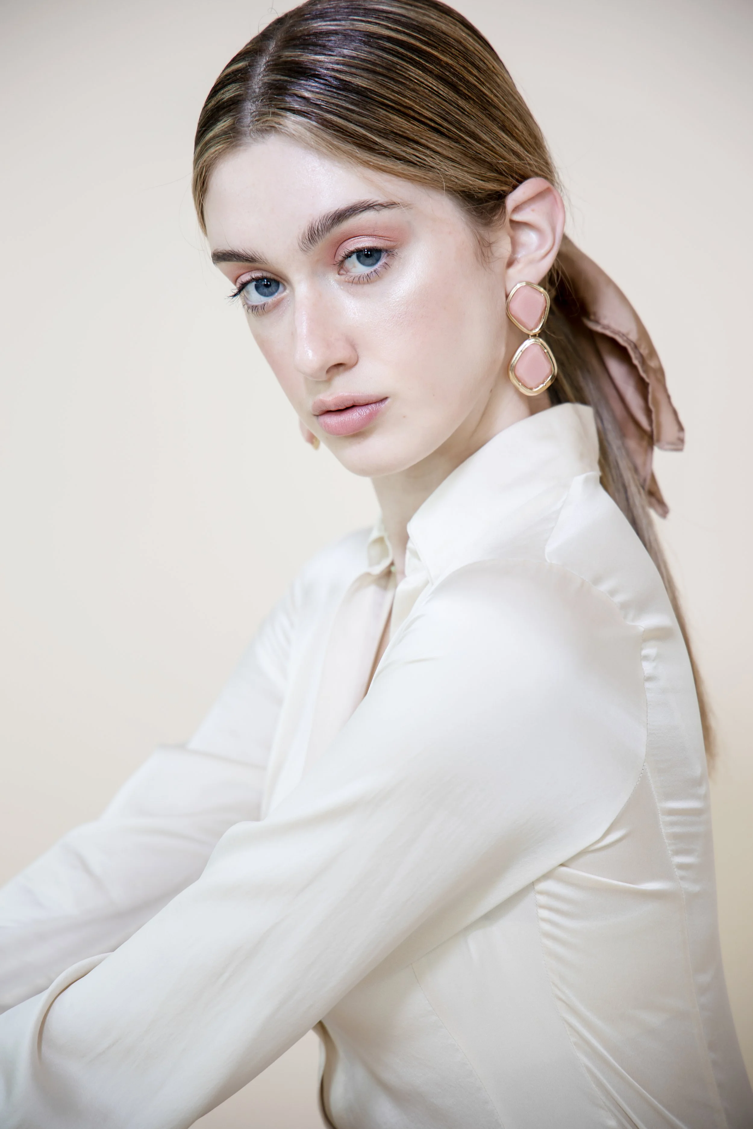 A woman with light skin and blue eyes, wearing a white satin blouse, pink earrings, and a pink neck scarf, is looking at the camera against a plain light background.