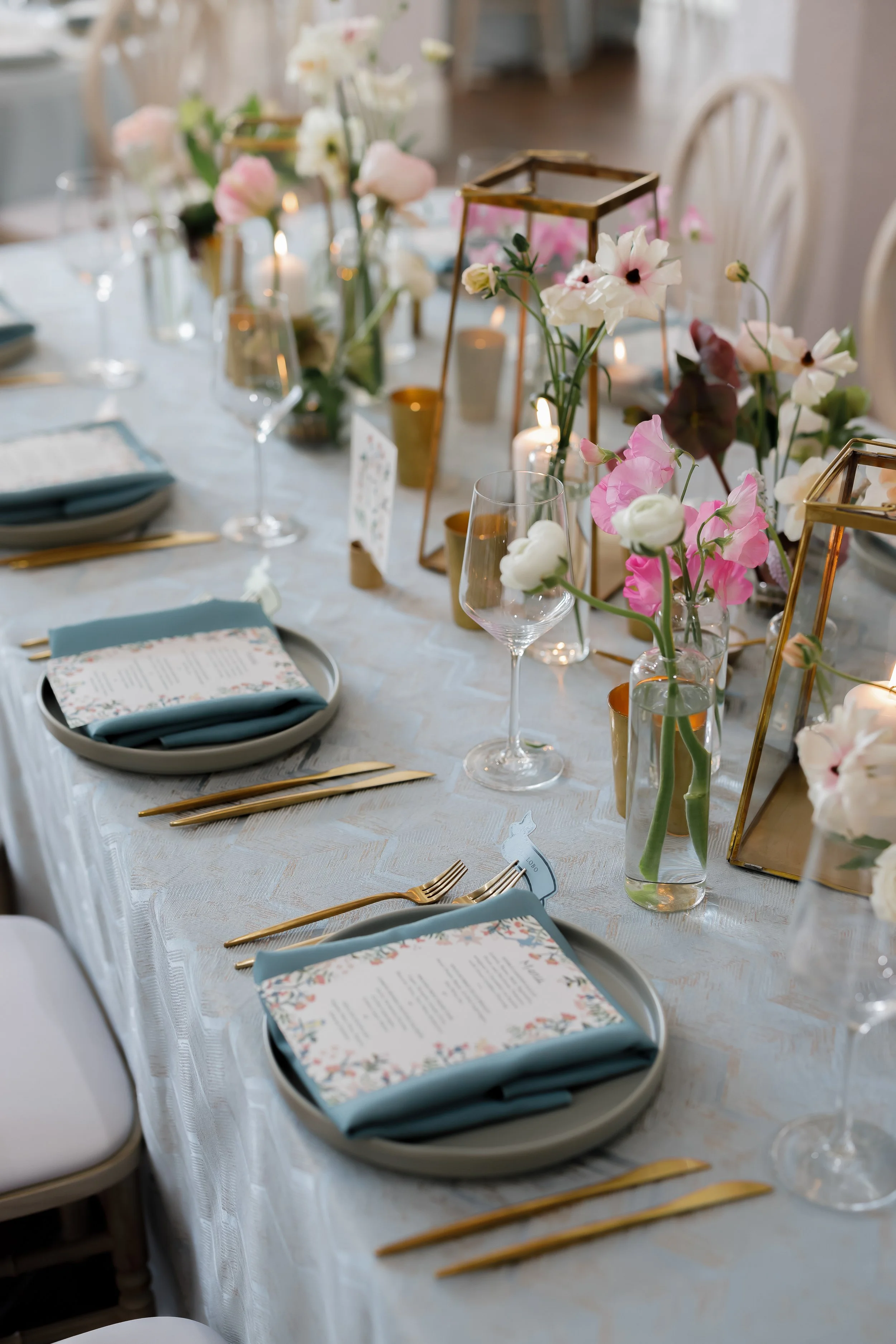 Decorated dining table with blue napkins, gold utensils, floral menus, vases with pink and white flowers, and glass candle holders, set for a formal event.