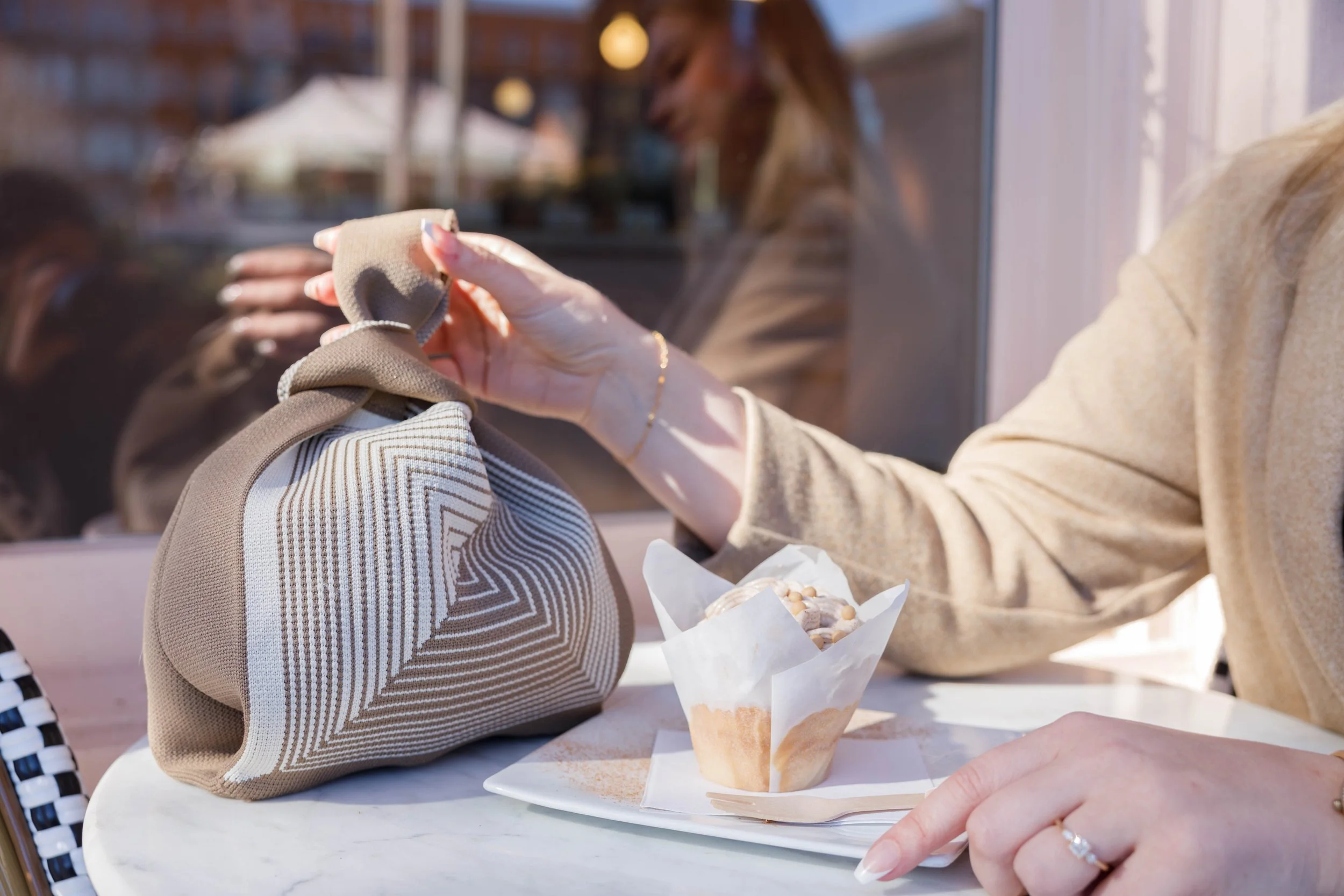 Person sitting at a table with a pastry, holding a beige bag, with another person visible through the window also inside the café.