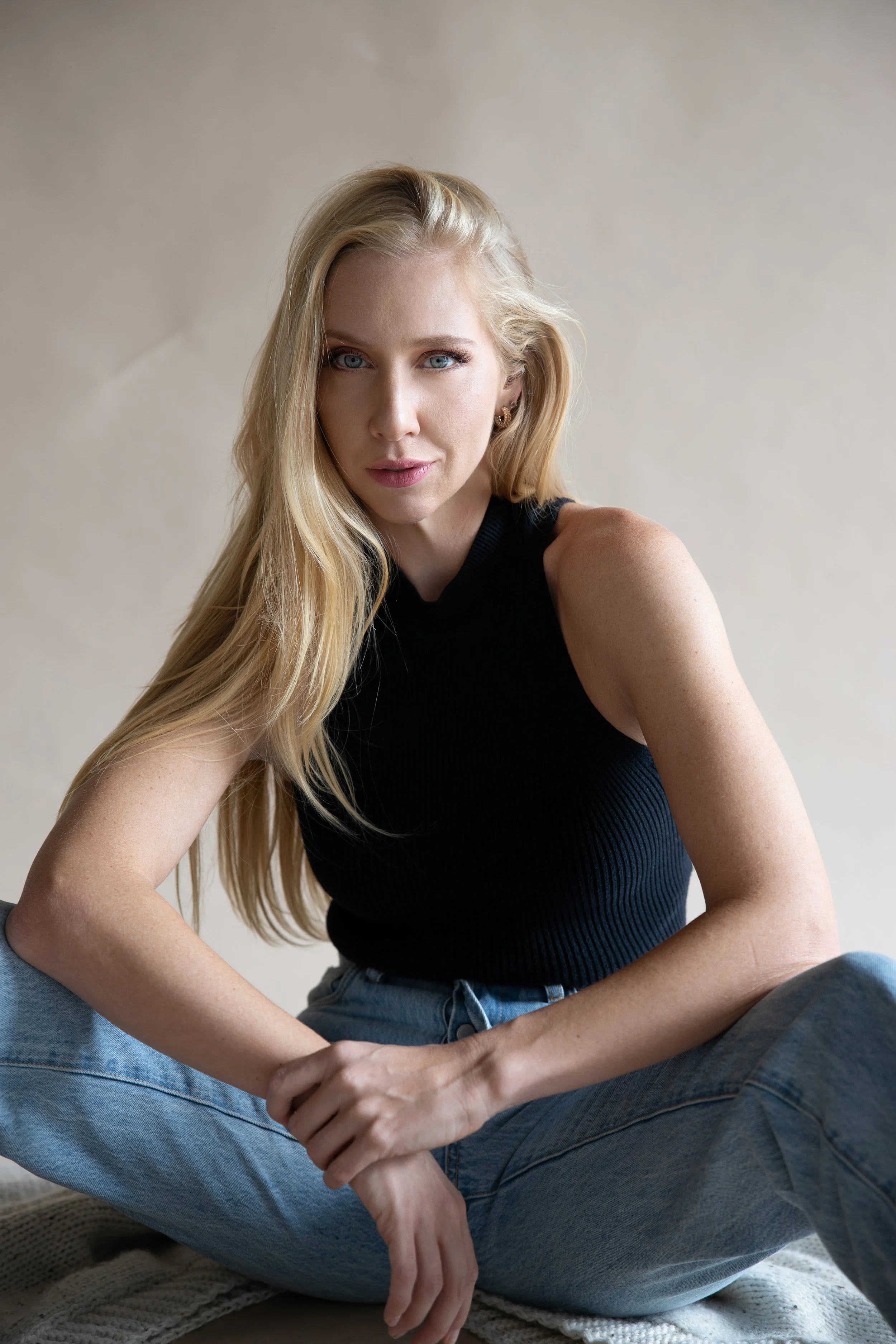 A young woman with long blonde hair, wearing a black sleeveless top and blue jeans, sitting cross-legged on a textured surface against a neutral background, looking directly at the camera.