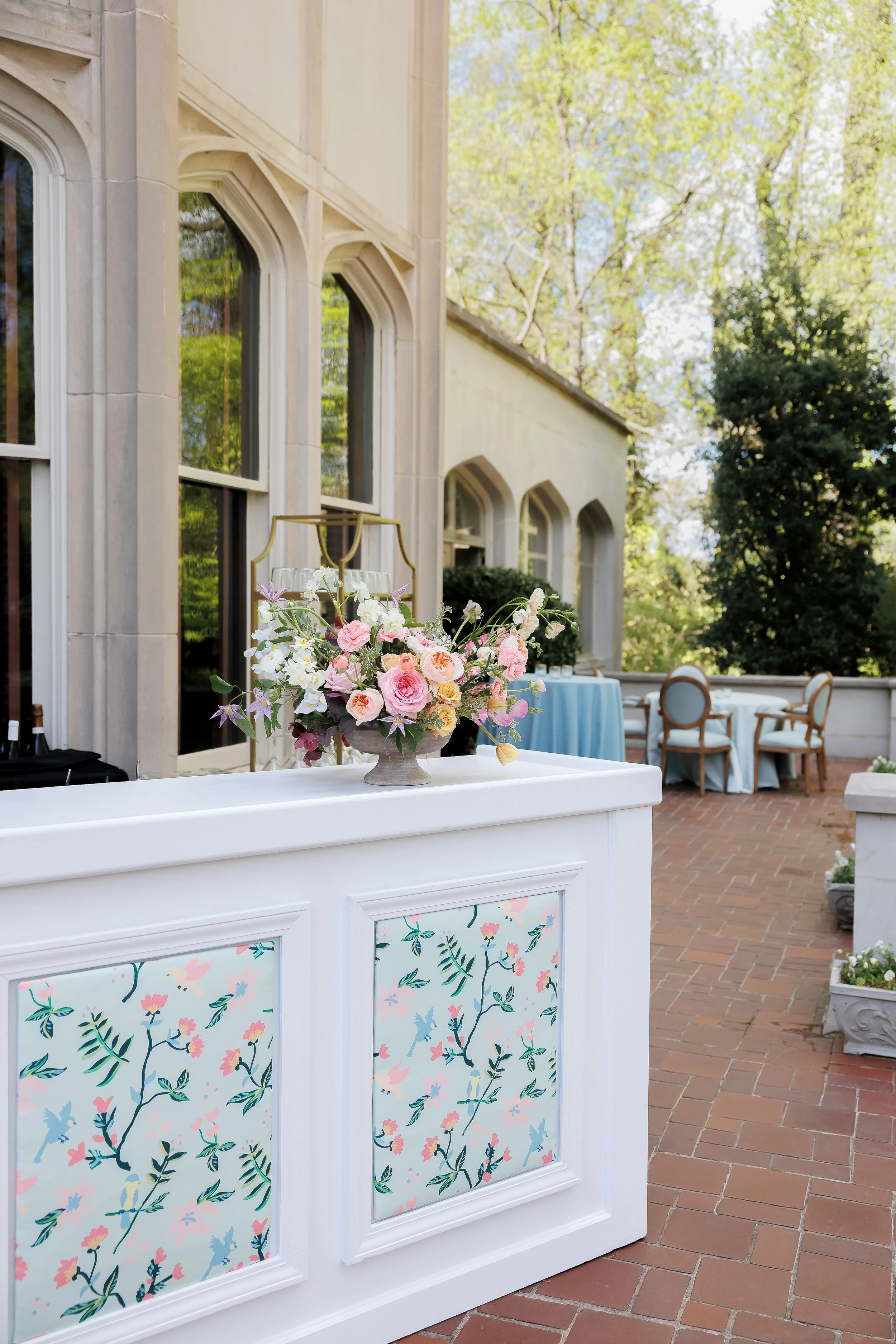 Elegant outdoor patio decorated for a special event, with a white floral arrangement, tables with blue tablecloths, and chairs surrounded by greenery.