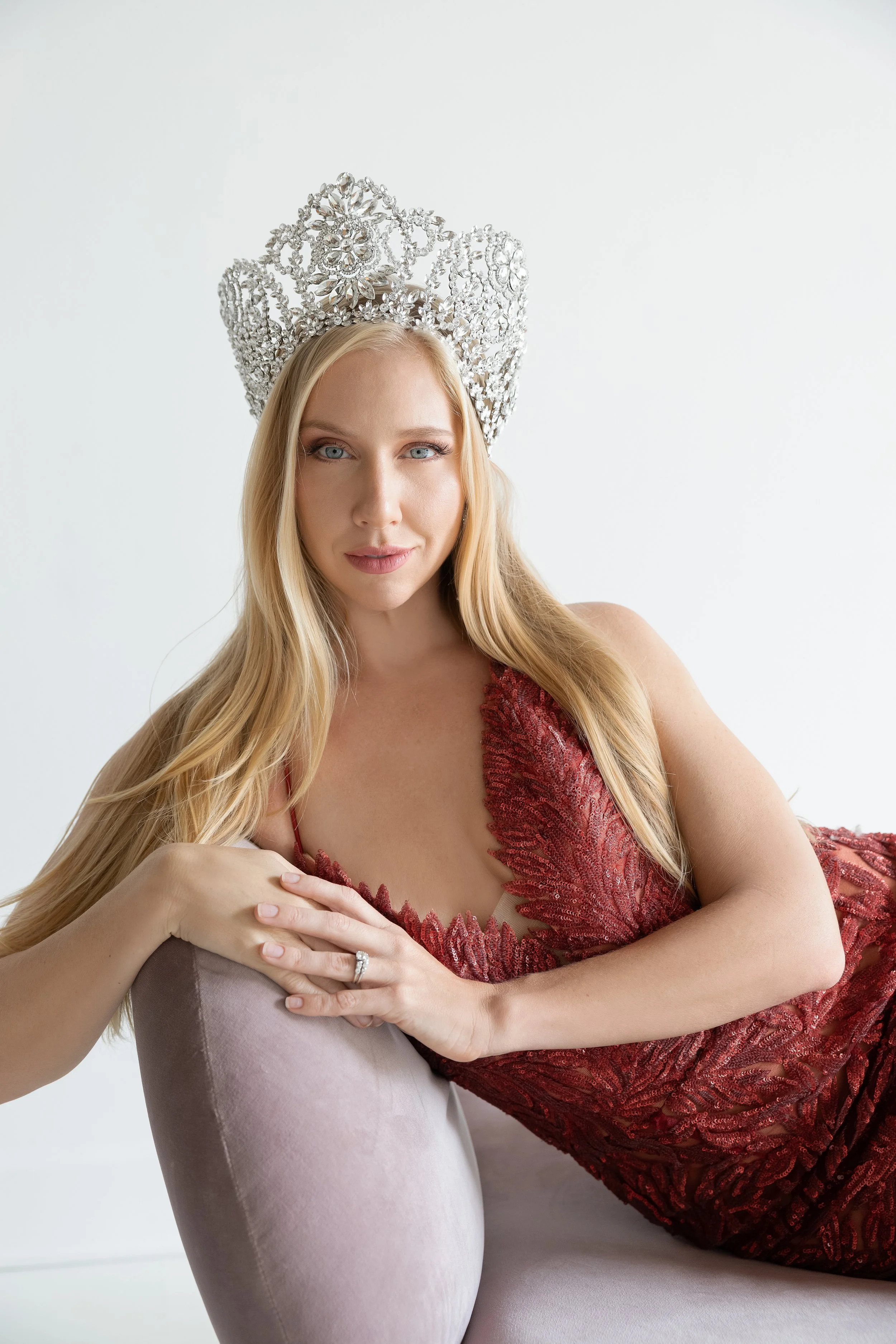 A woman with long blonde hair and blue eyes wears a large, ornate silver crown and a red lace dress, and poses reclining on a light purple armchair against a plain white background.