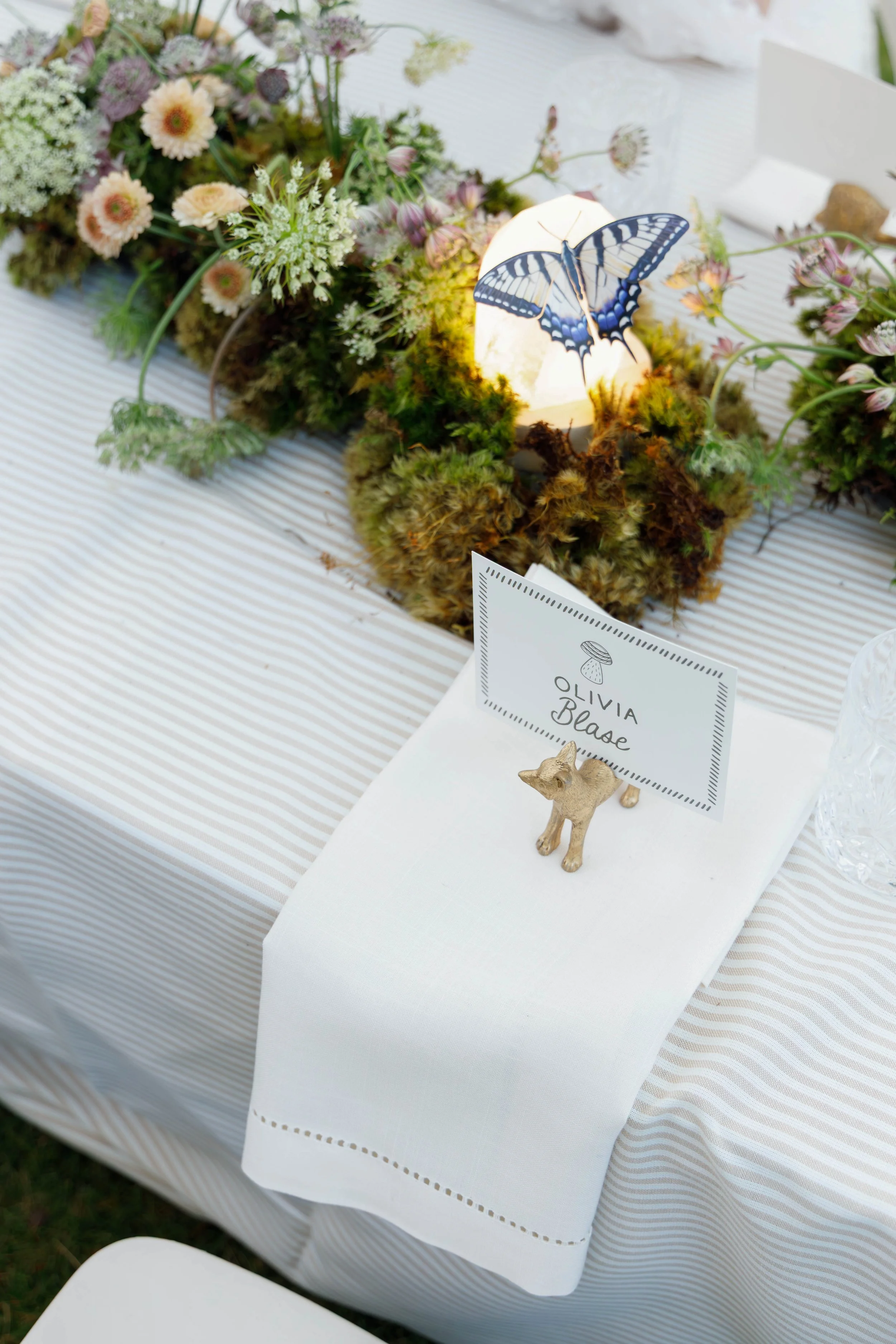 A table decorated with a floral centerpiece, a white napkin, a name card for Olivia Blease, and a small animal figurine, with a butterfly decoration and a glowing light in the background.