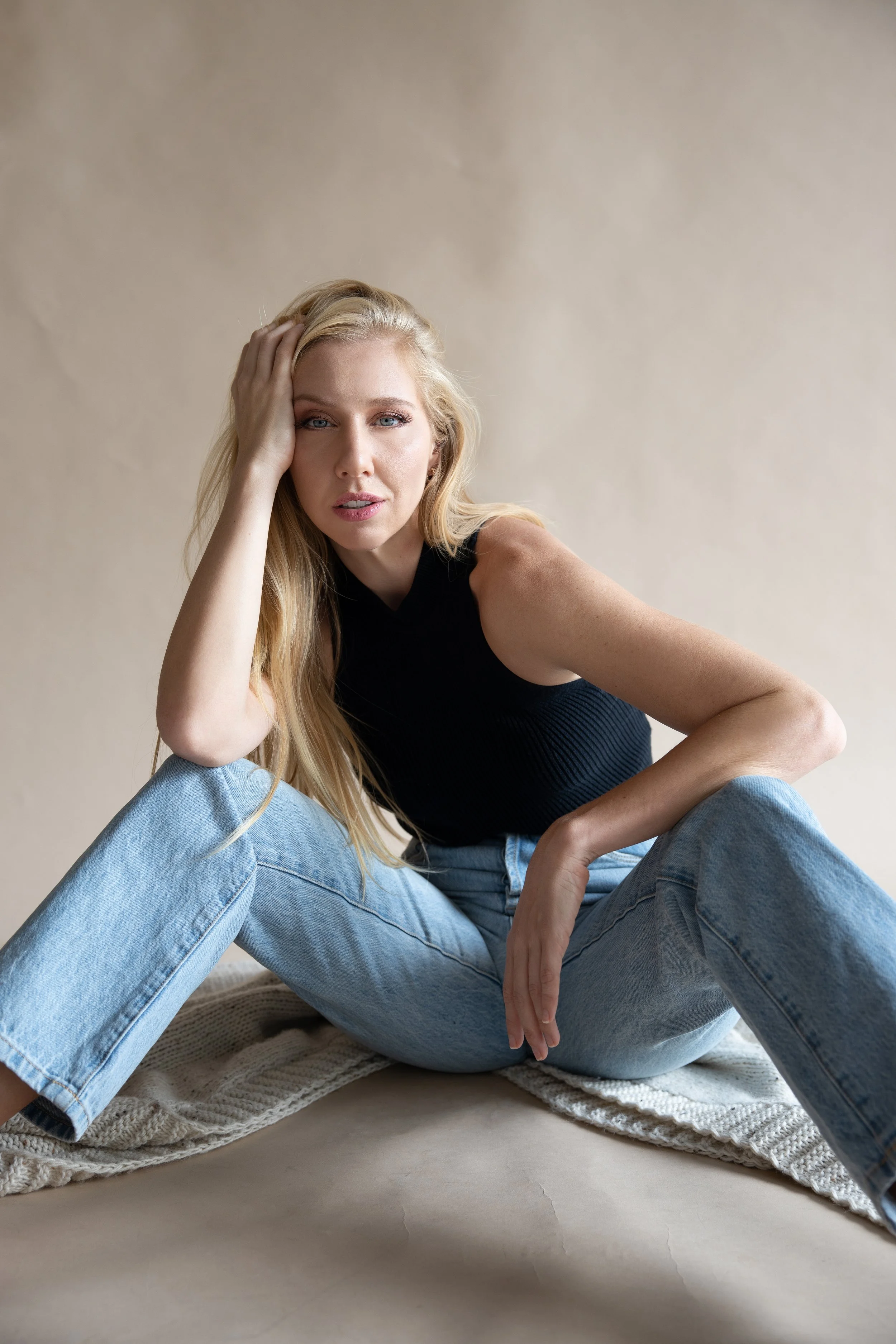 A woman with long blonde hair sitting on a carpeted floor, wearing a black sleeveless top and light blue jeans, with one hand resting on her head and looking at the camera.
