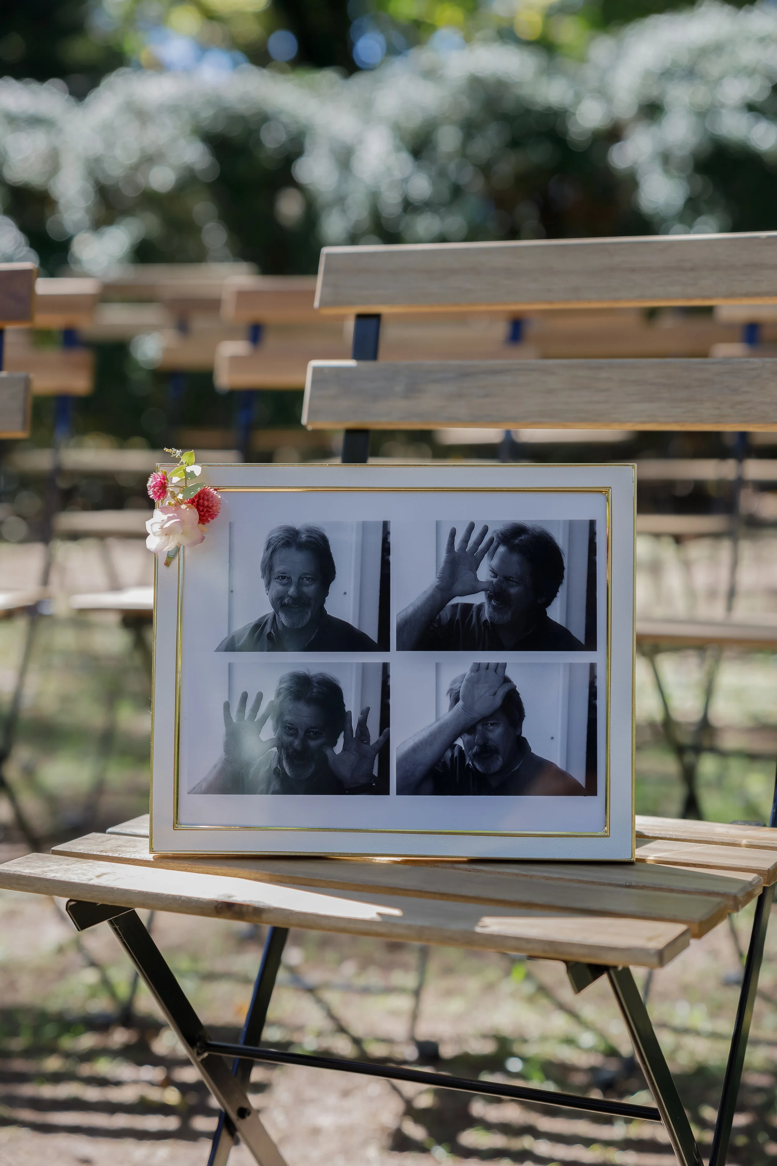A framed collage of four black and white photos of a man with a beard, each showing different playful facial expressions and gestures, resting on a wooden folding table outdoors with empty wooden benches and greenery in the background.