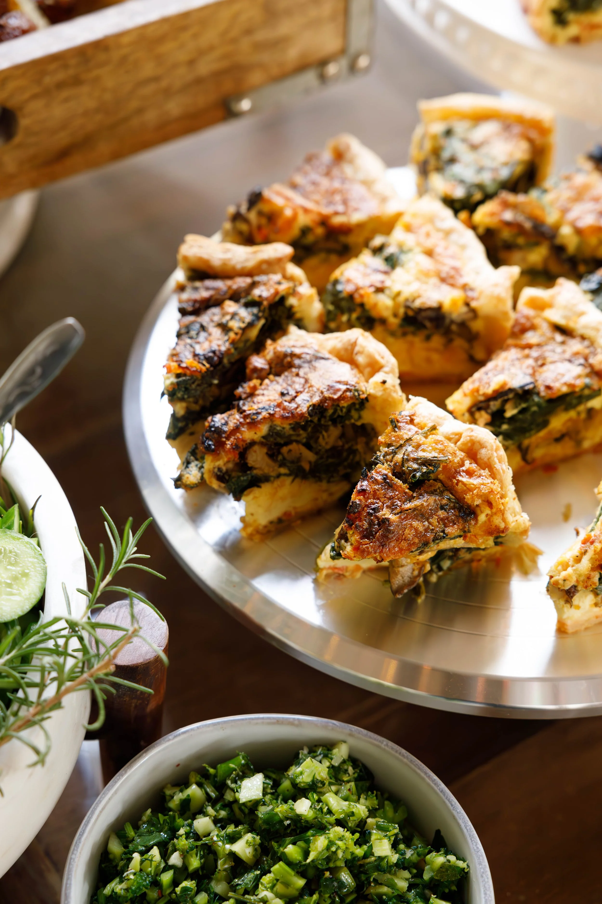 Slices of spinach quiche on a silver platter with a side of chopped green salad and a bowl of fresh herbs and cucumber slices.