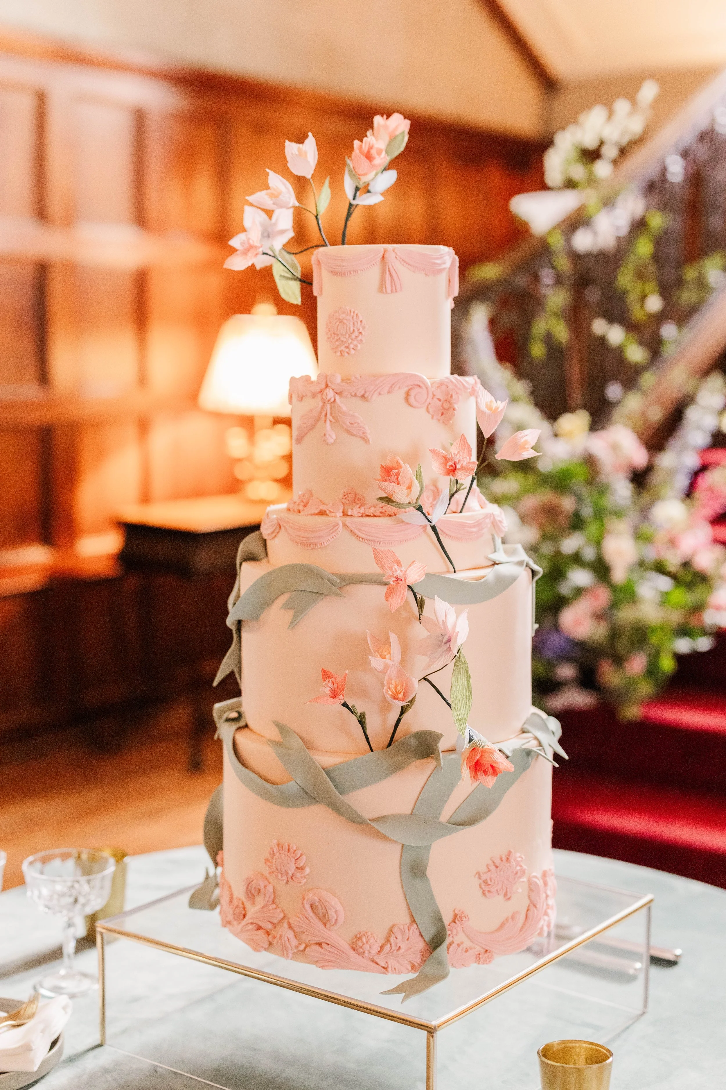A four-tier pink wedding cake decorated with pink and green sugar flowers and gray ribbons, set on a stand with a gold frame, in a decorated reception hall.