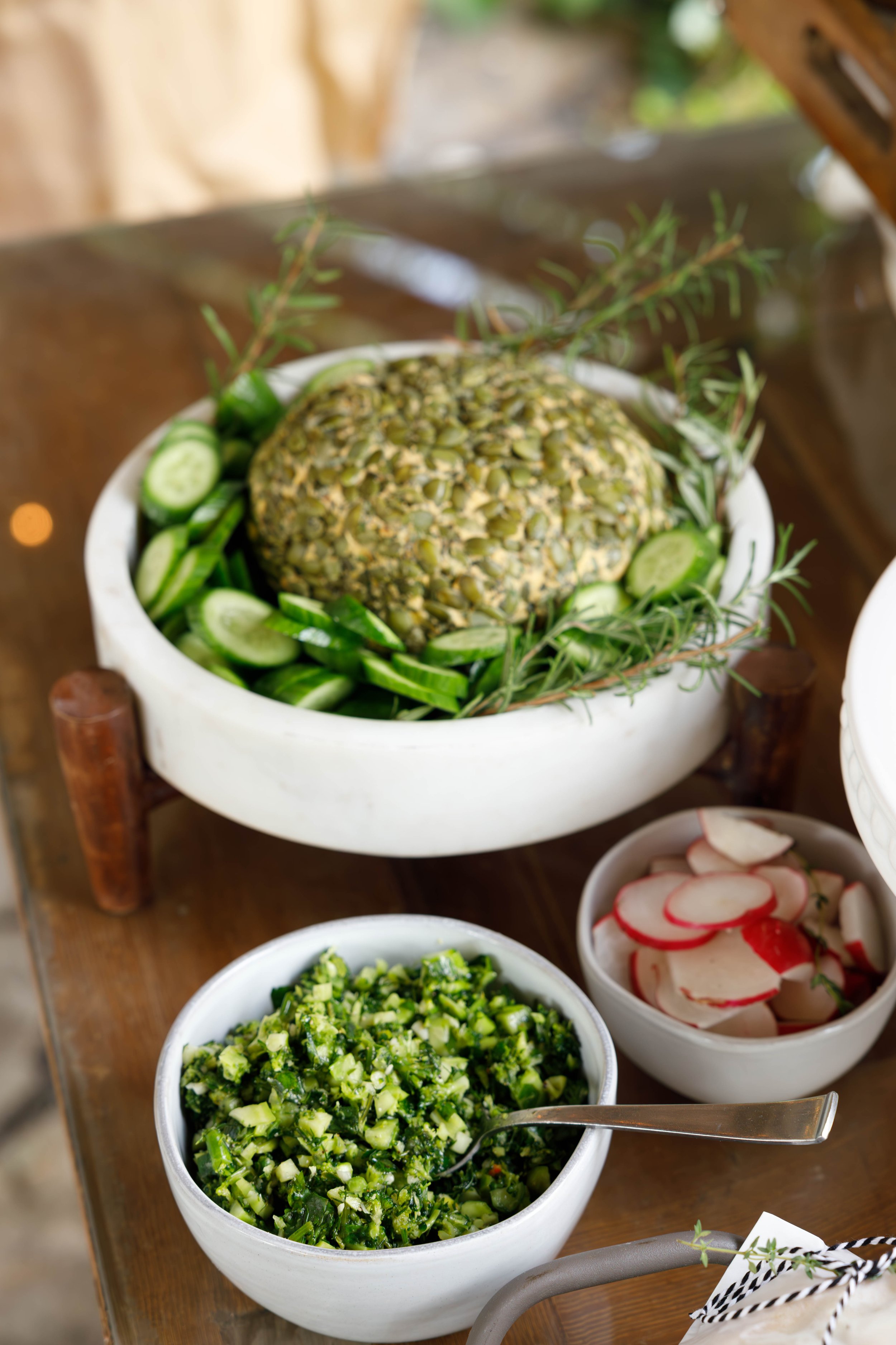 A large bowl with a pineapple surrounded by sliced cucumbers and sprigs of herbs, and two smaller bowls of chopped vegetables on a wooden table.