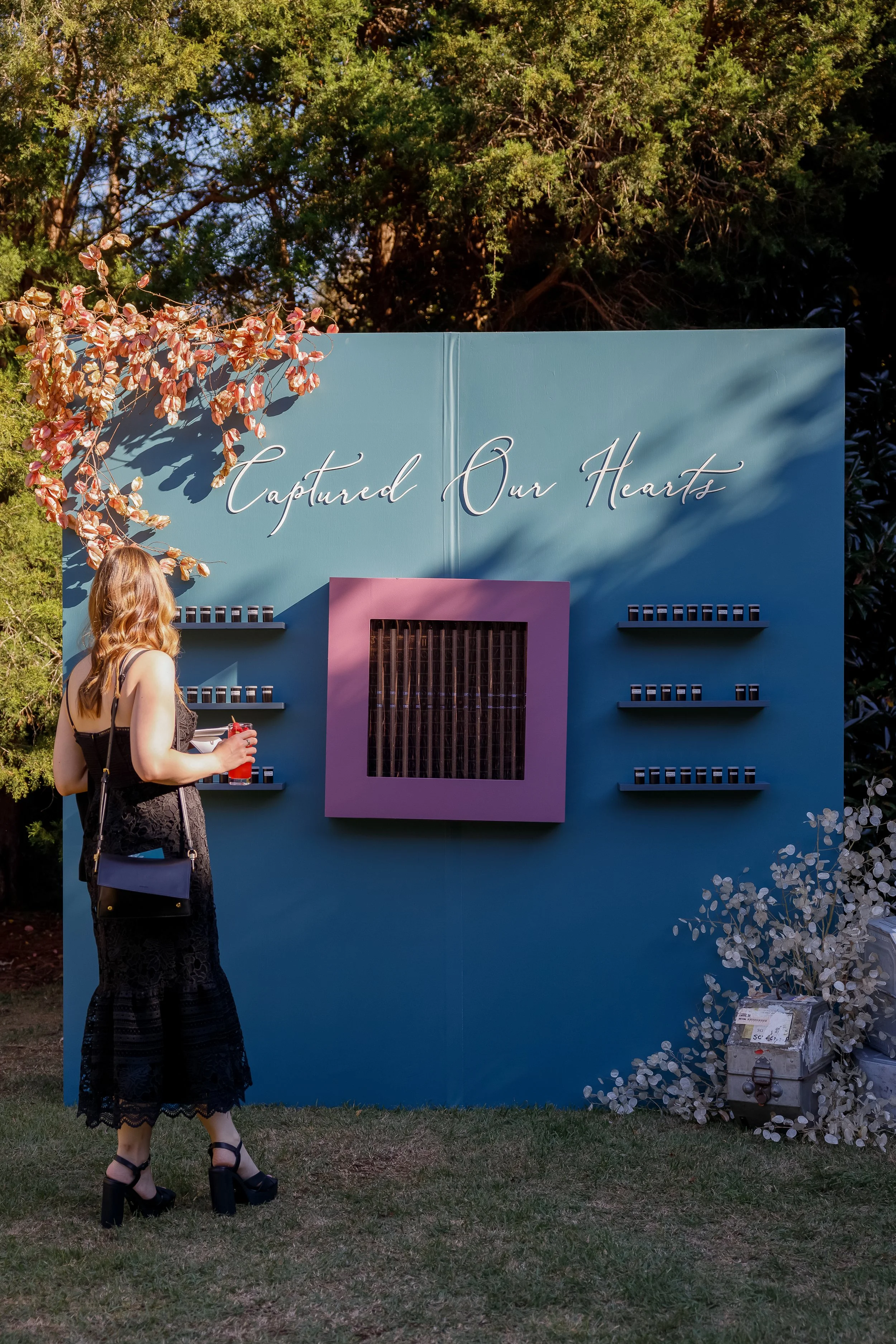 A woman standing outdoors in front of a blue display with the words 'Captured Our Hearts' written in cursive. She is holding a red drink, wearing a black lace dress and black high heels, and has a black purse hanging from her shoulder.