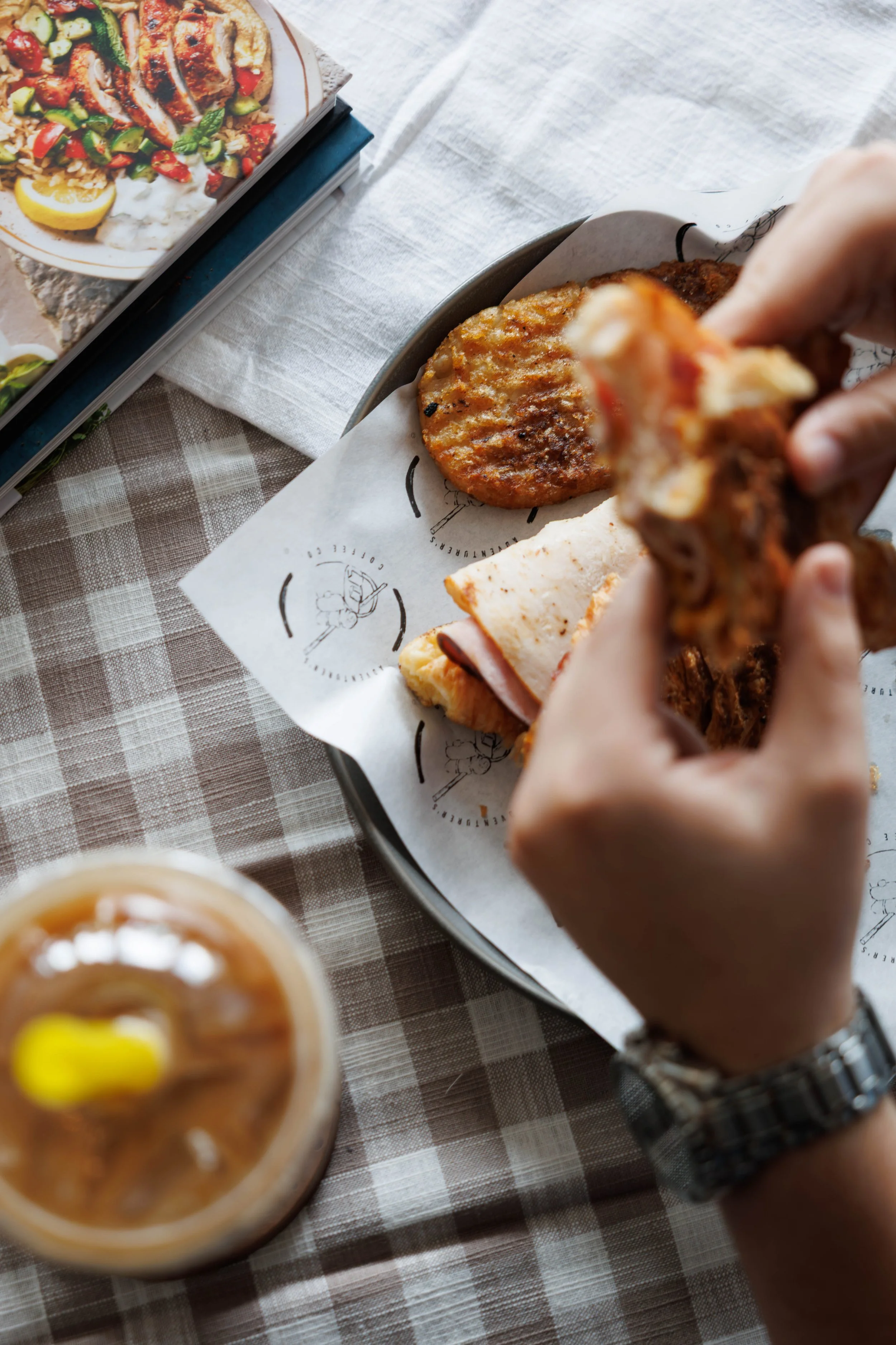 Person holding a half-eaten sandwich with potato cakes on a serving tray, a drink with a lemon wedge, and a cookbook on a checkered tablecloth.