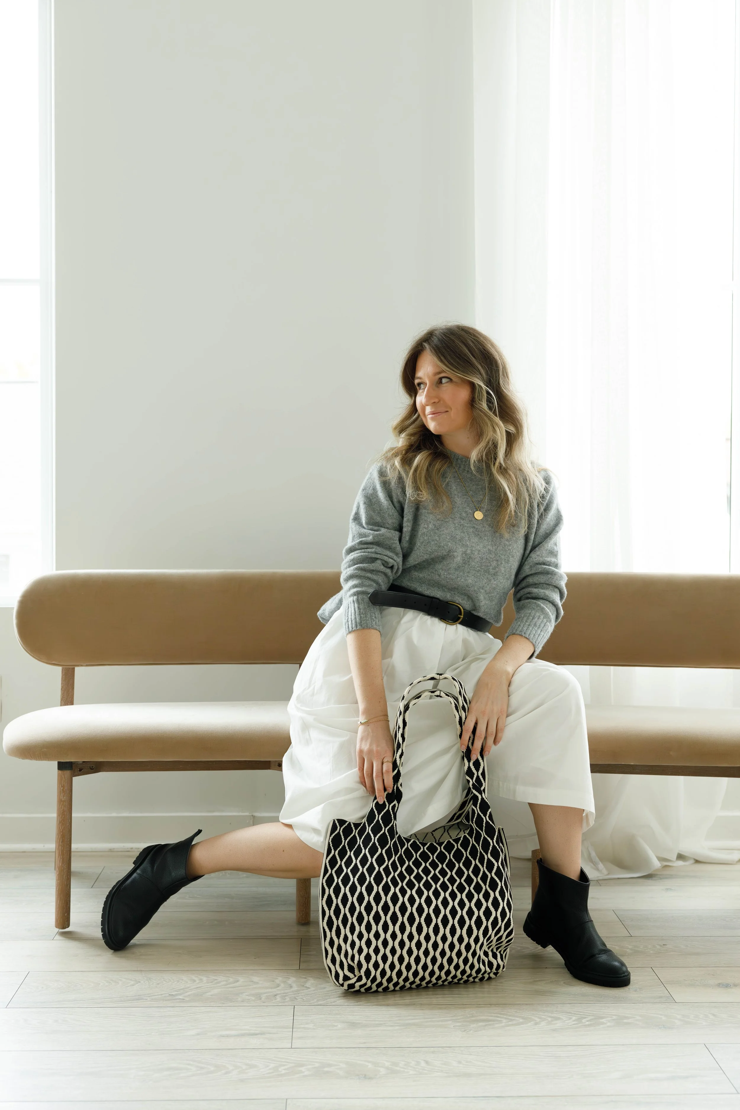 A woman with wavy, shoulder-length hair sitting on a beige bench in front of a white wall and large window. She is wearing a gray sweater, white wide-leg pants with a black belt, black ankle boots, and holding a black-and-white patterned tote bag. Sh