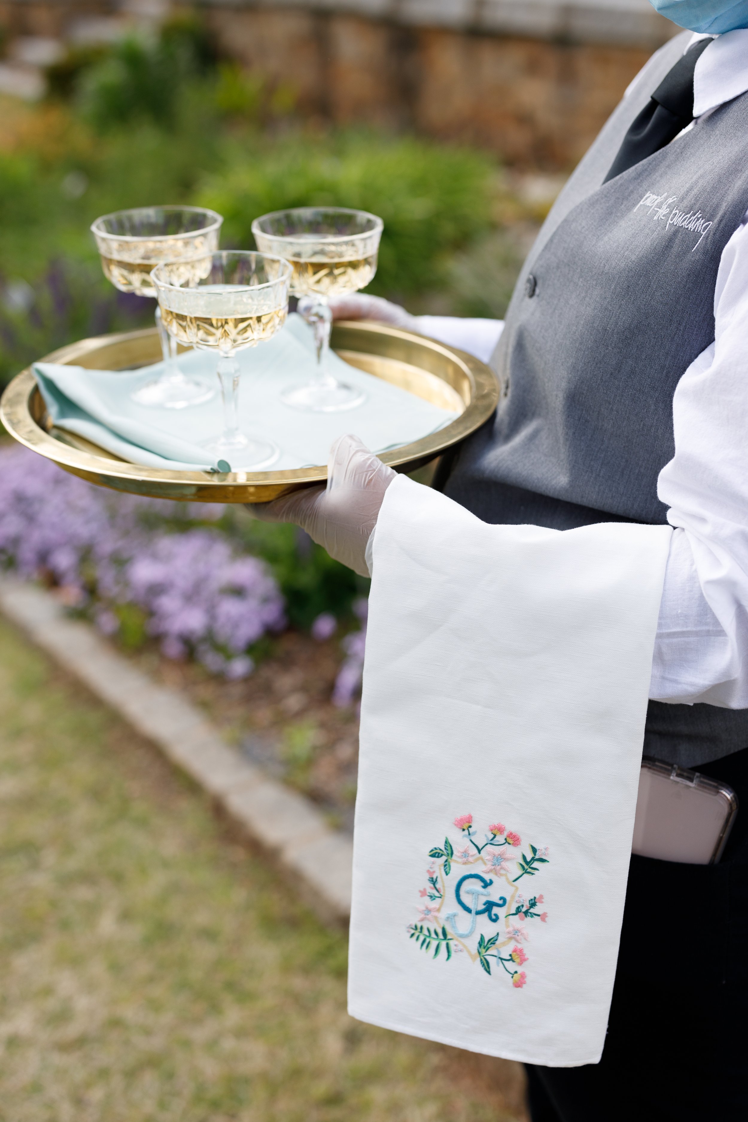 A person dressed in formal attire holding a tray with four glasses of white wine outdoors, with a garden and flowers in the background.