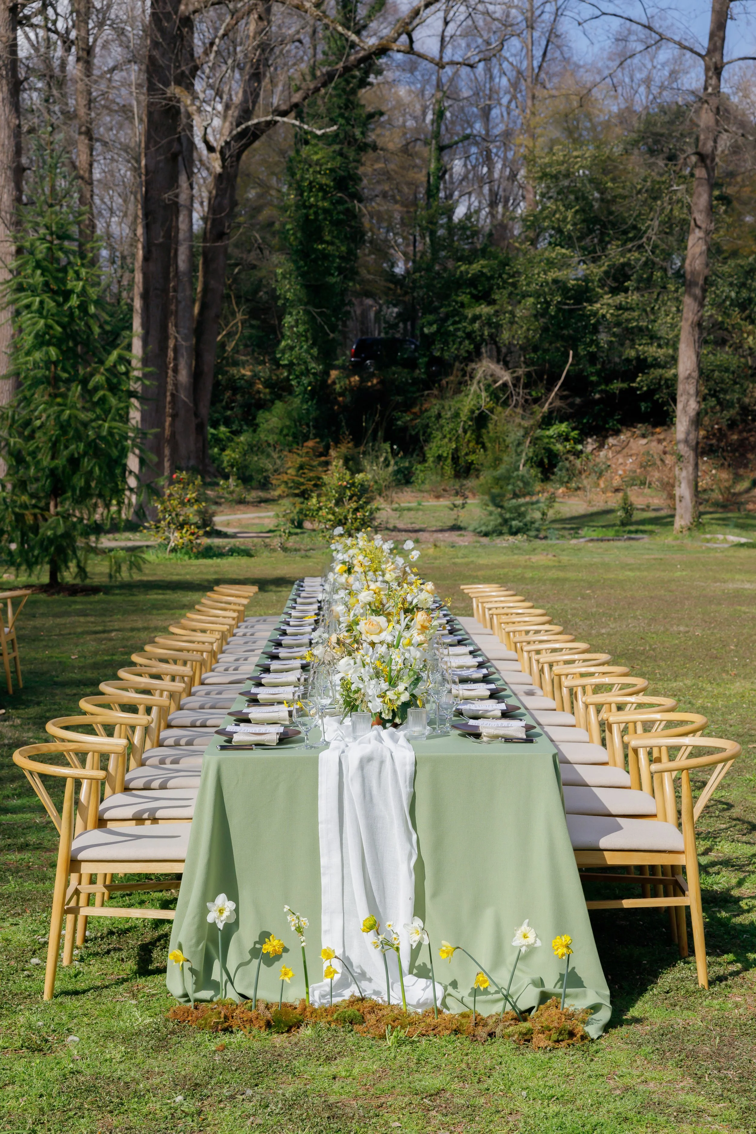 Outdoor wedding reception setup with a long rectangular table covered in a green tablecloth, decorated with flower arrangements, table settings, and surrounded by wooden chairs with white cushions, set in a lush garden with trees and greenery in the 