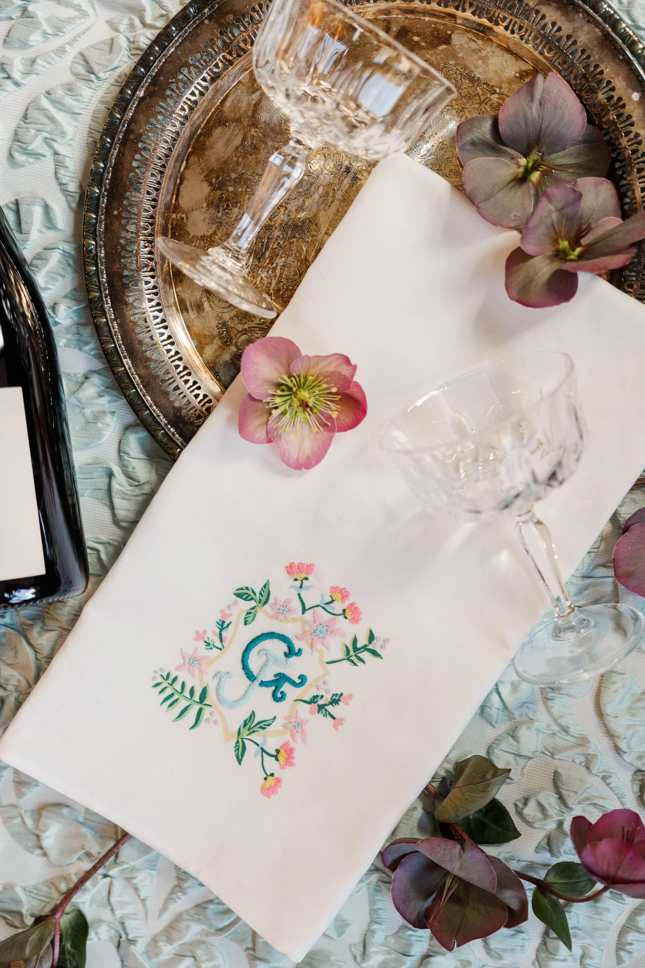 A decorative table setting featuring two empty crystal wine glasses on a white embroidered napkin with a floral monogram, placed on a silver tray decorated with pink flowers and dark green leaves, with additional pink and green floral accents around.