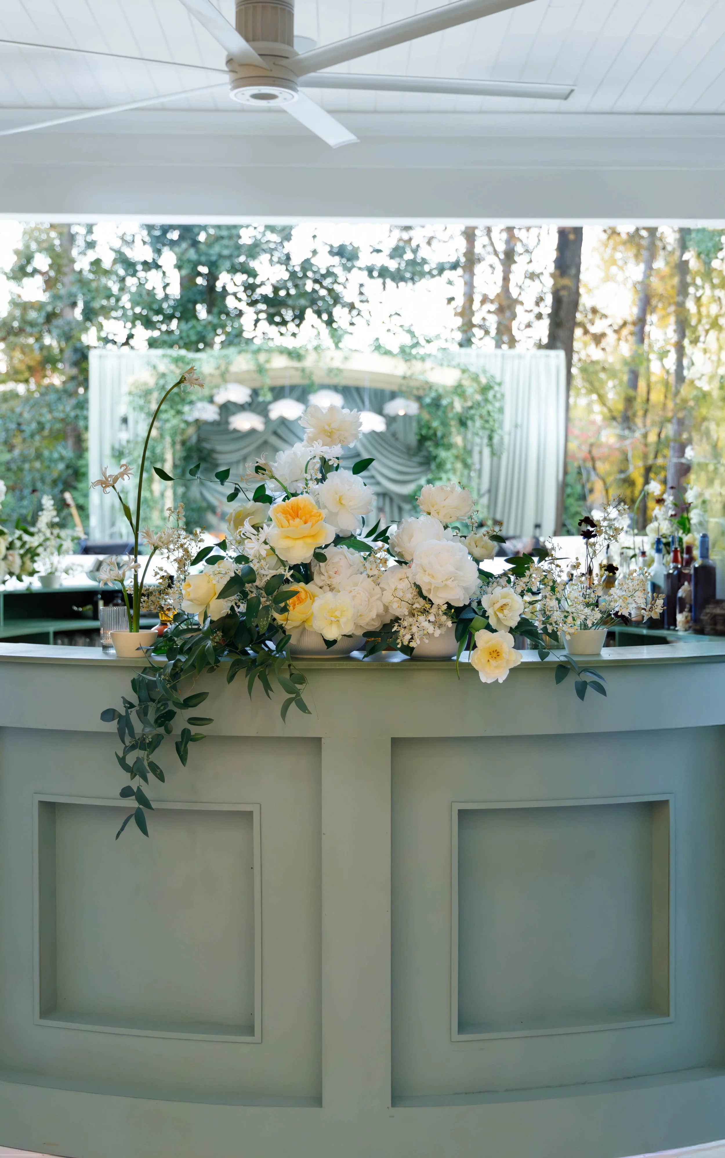 A bar decorated with a large arrangement of white and pale yellow flowers and greenery, with a background of trees and a window. There are bottles and barware on the right side of the bar.