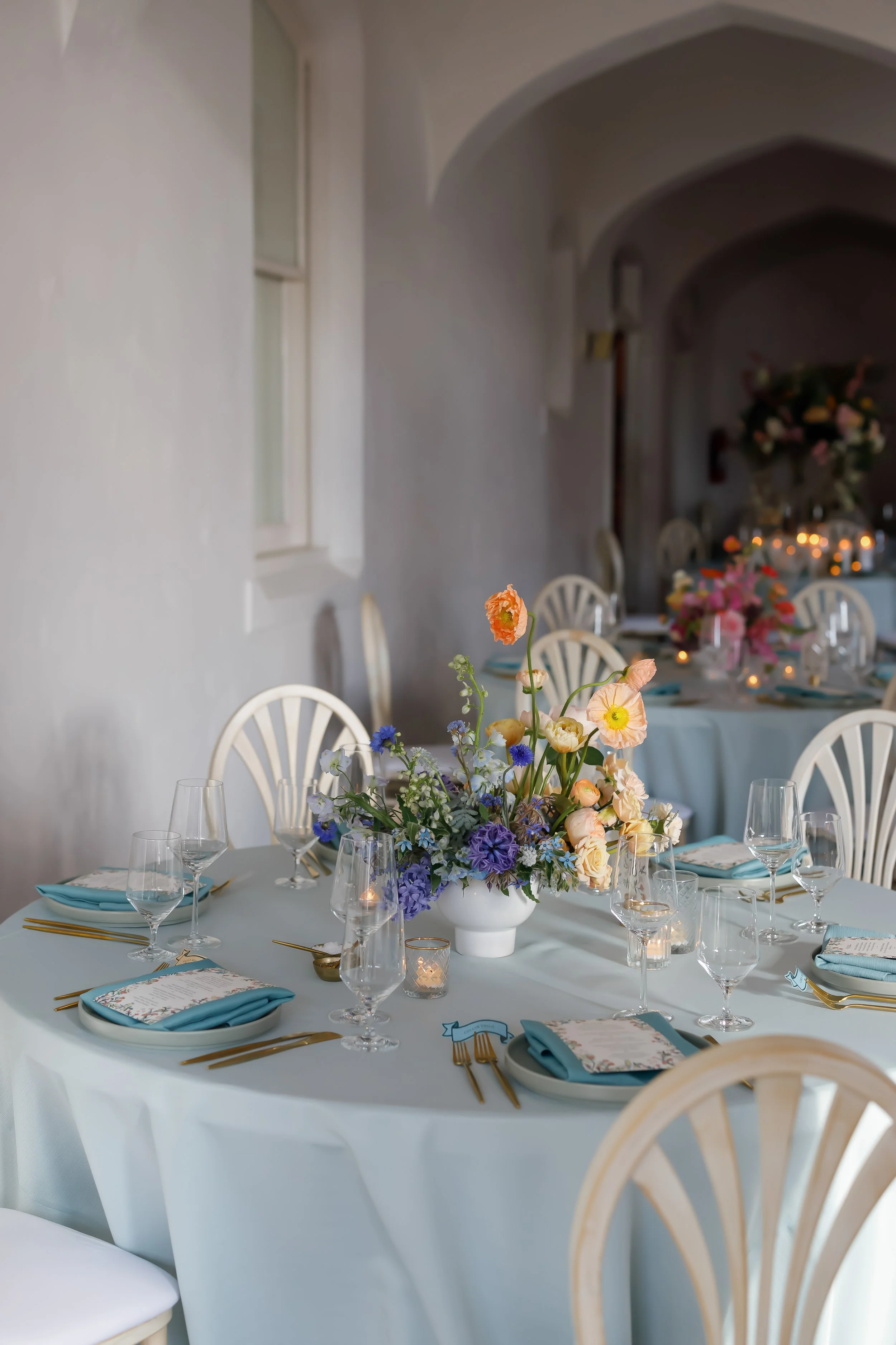 A round banquet table decorated with a pastel blue tablecloth, set for a formal event with gold silverware, blue napkins, wine glasses, and a floral centerpiece featuring pastel and purple flowers. The background shows additional tables with similar 