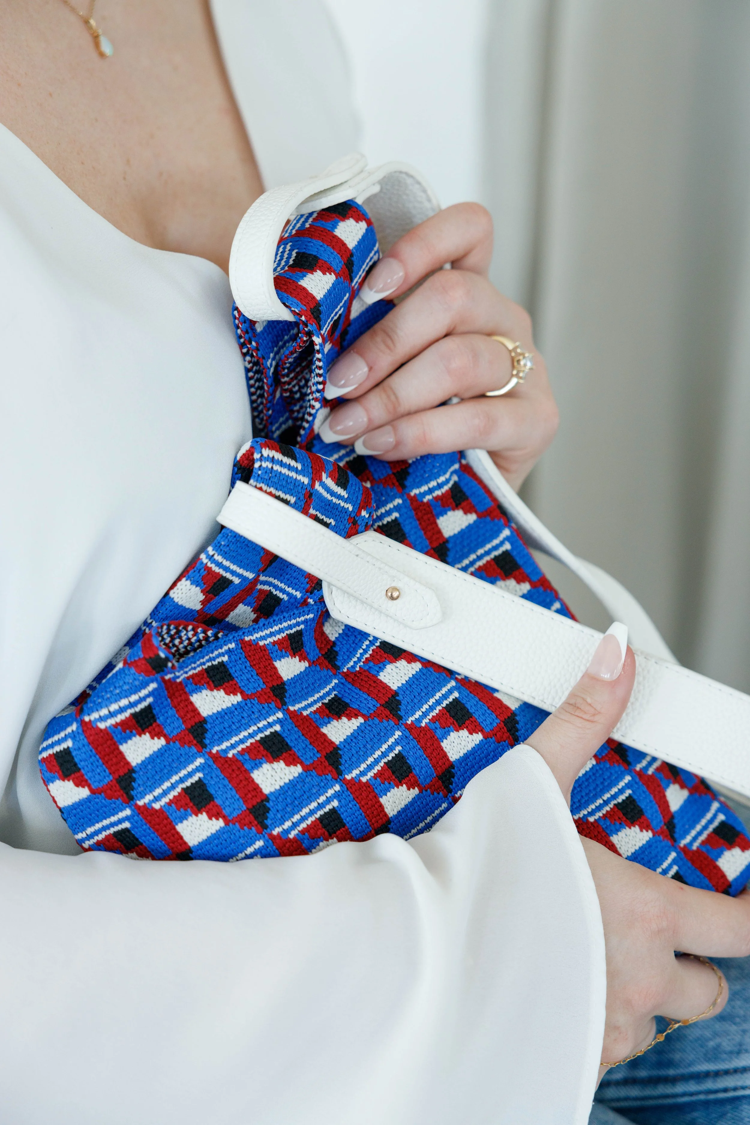 Person holding a patterned fabric bag with a white strap, wearing a white long-sleeve top and a gold ring.