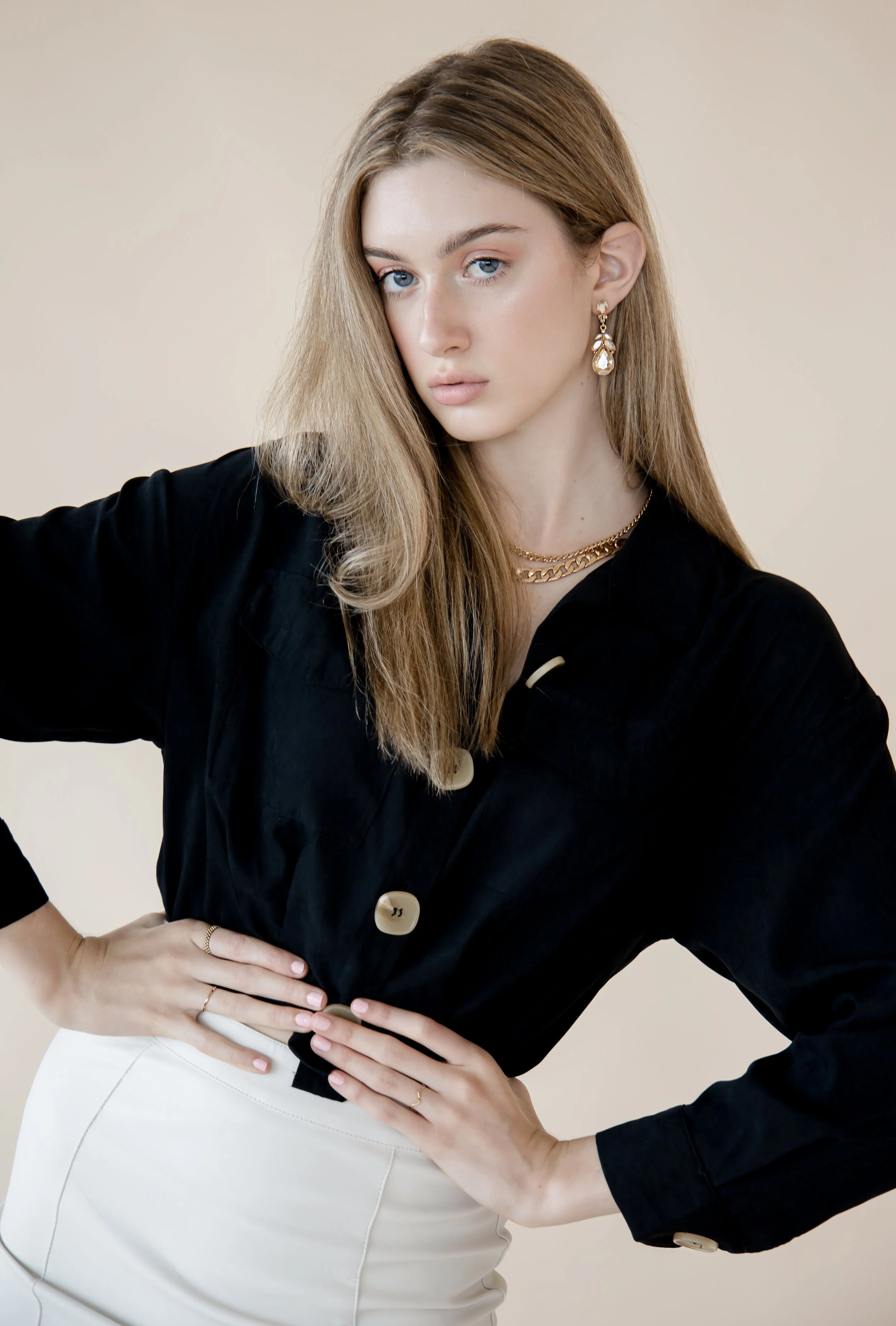 A young woman with long, wavy blonde hair and blue eyes posing with her hands on her hips against a beige background. She is wearing a black button-up shirt, white pants, and gold jewelry, including earrings, necklaces, and rings.