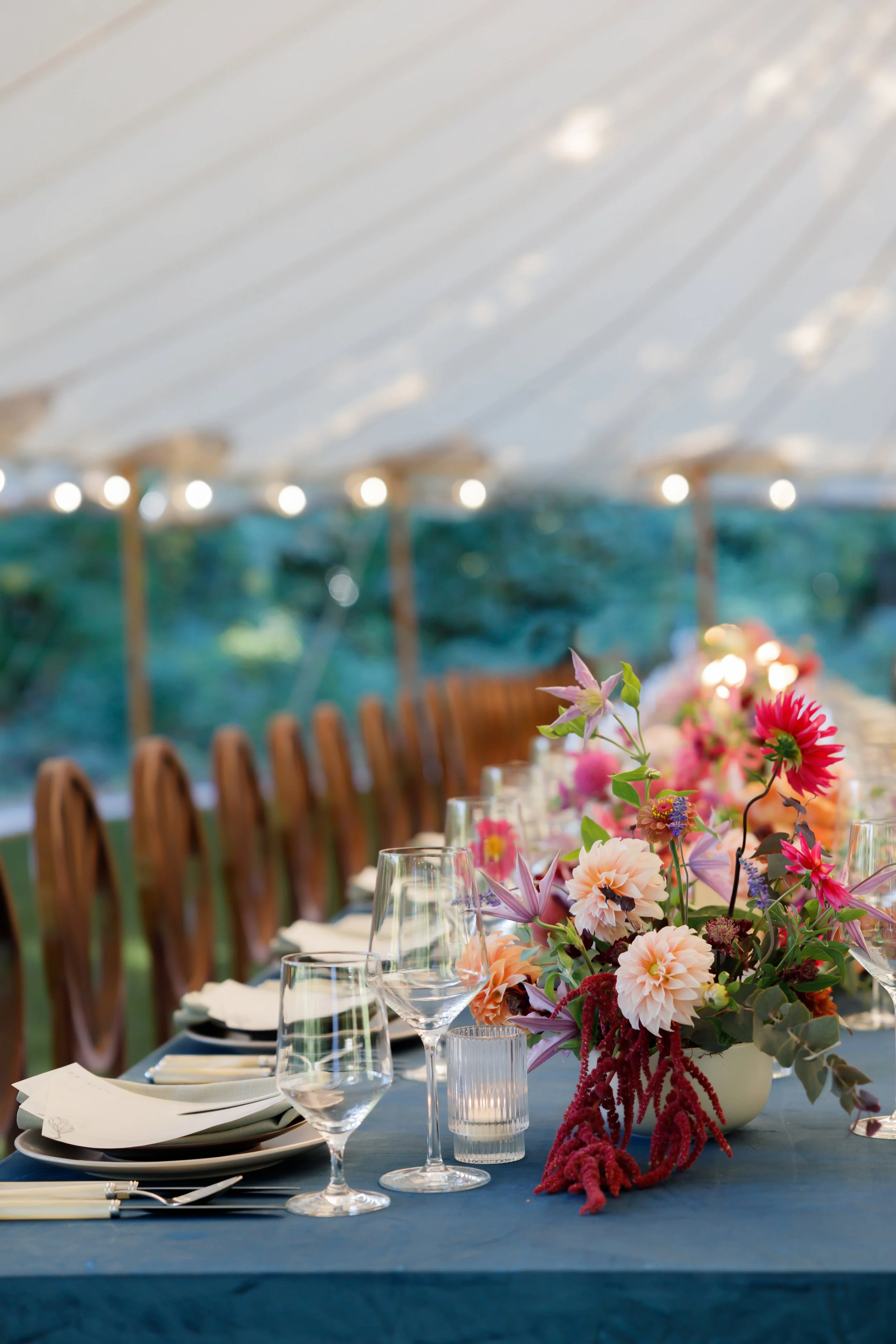 Elegant outdoor dining table set with glassware, napkins, and a colorful floral centerpiece under a canopy.