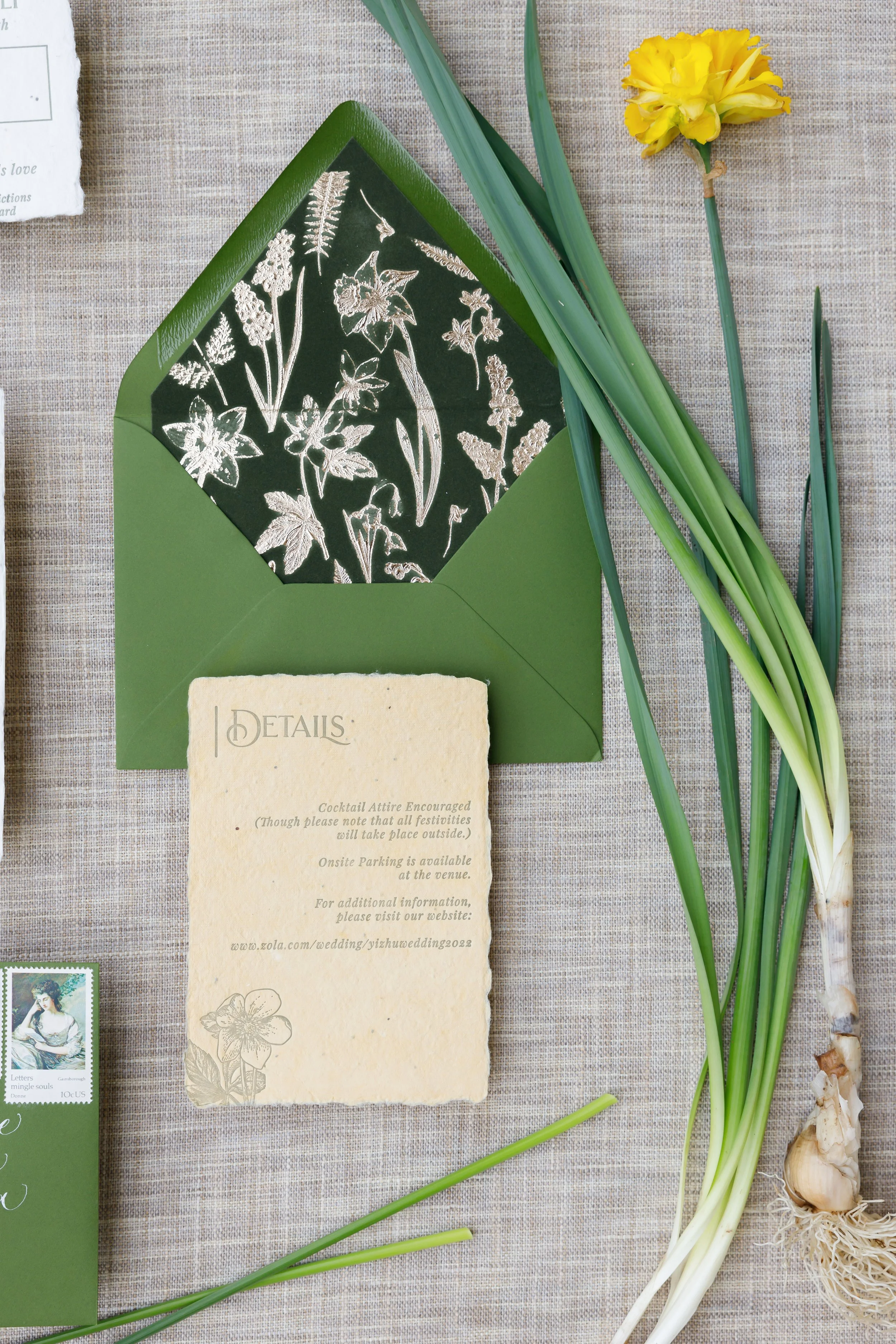 A wedding invitation layout with a green envelope containing a floral-patterned card, accompanied by a bunch of green onions and a yellow flower on a textured beige surface.
