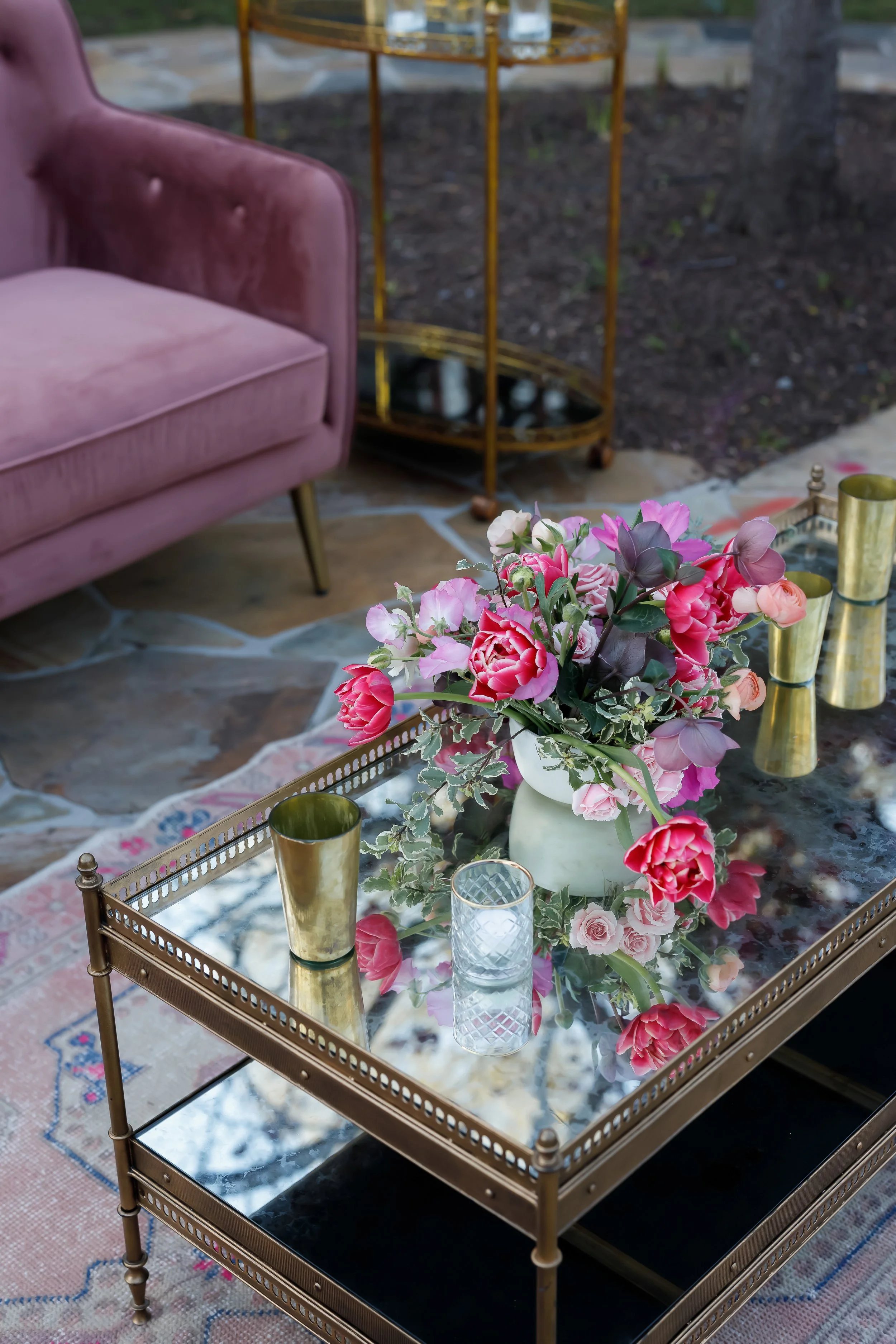 A glass-top coffee table with a floral arrangement in a white vase, surrounded by gold-colored candle holders, placed on a pink and blue patterned rug. A pink velvet sofa and a gold shelf are visible in the background.