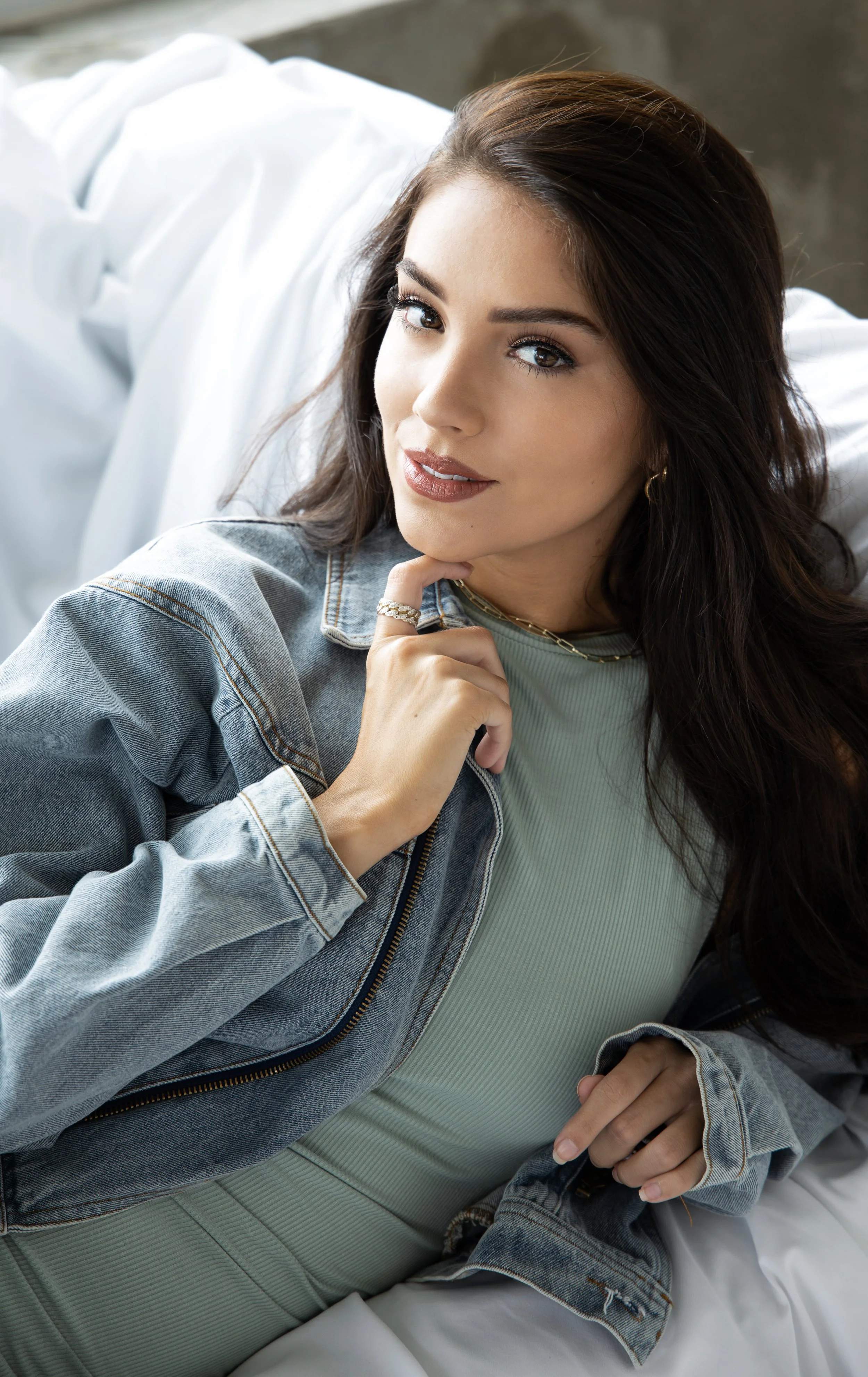 A young woman with long dark hair, wearing a denim jacket and light green top, is lying down on a bed, looking at the camera with a thoughtful expression, resting her chin on her hand.