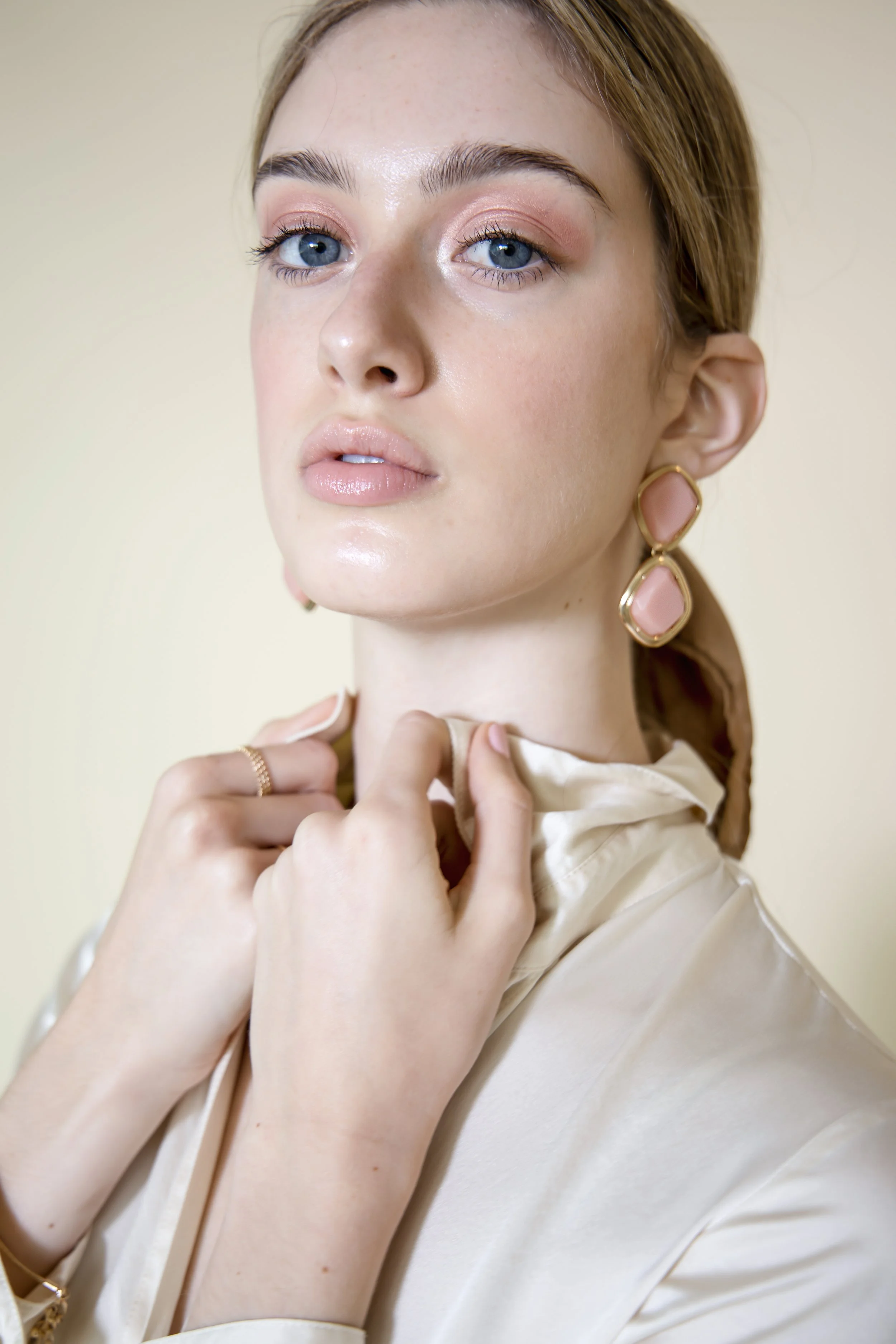 Close-up portrait of a young woman with fair skin, blue eyes, and blonde hair tied back. She is wearing pink eye makeup, glossy light pink lips, and gold earrings with pink stones. She has a gentle expression and is holding the collar of her light-co