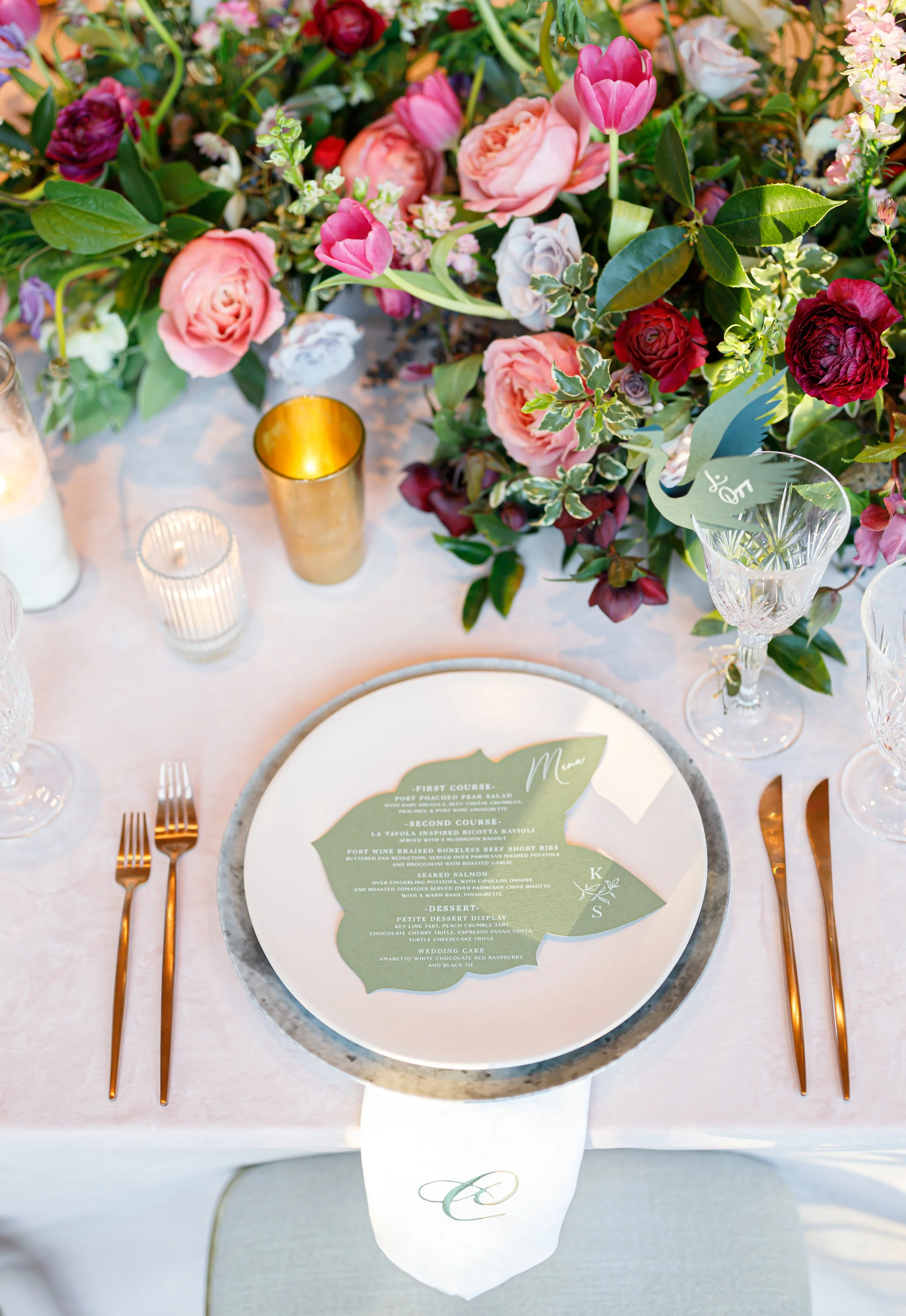Elegant table setting with a floral centerpiece of pink, red, and purple flowers, gold and glass candles, crystal glassware, and a printed menu on a green leaf-shaped card.