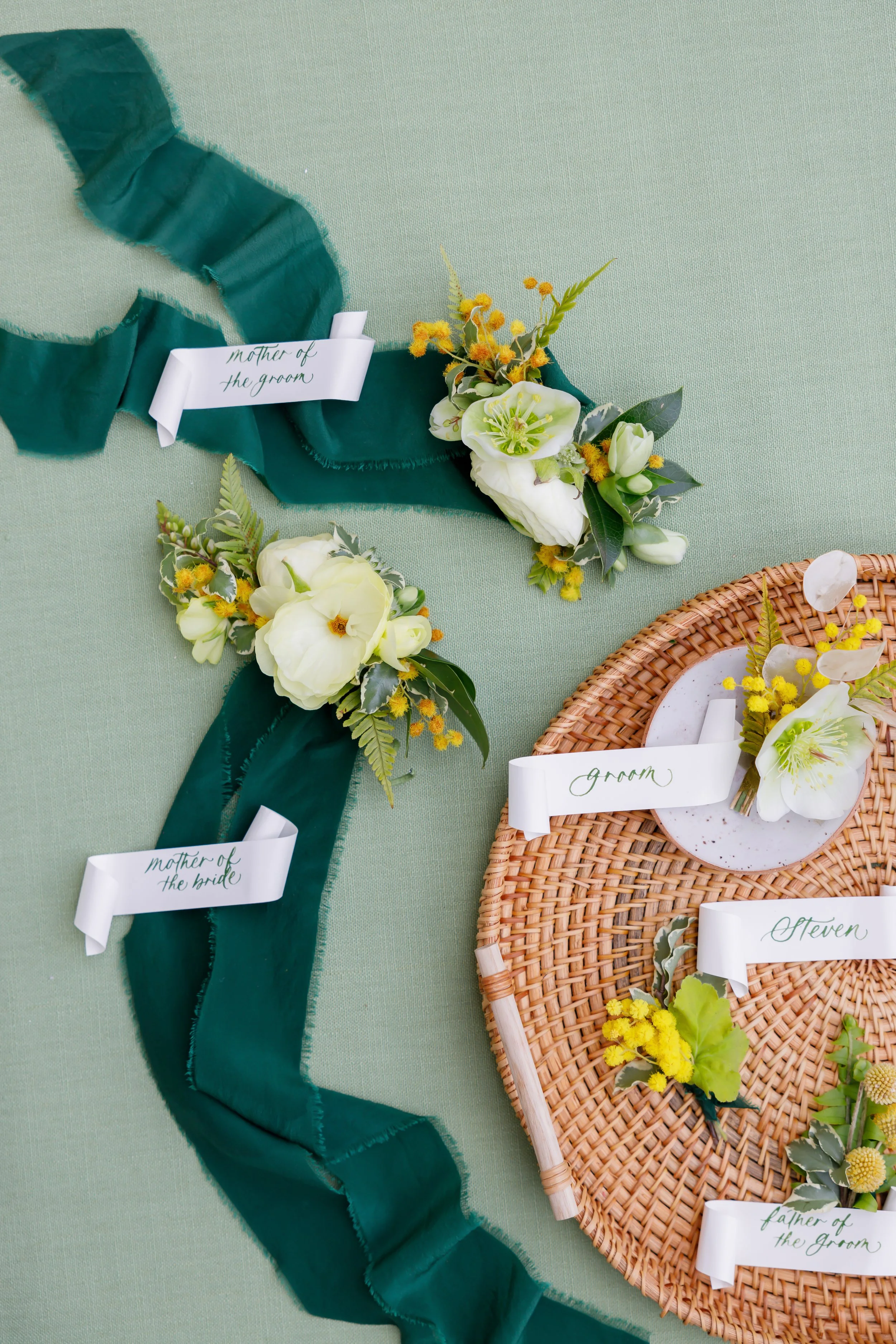 Floral corsage and boutonnieres with labels for a wedding, on a green tablecloth. The corsage has a tag labeled 'mother of the groom' and the boutonniere has tags labeled 'groom,' 'bride,' 'father of the groom,' and 'seven.'