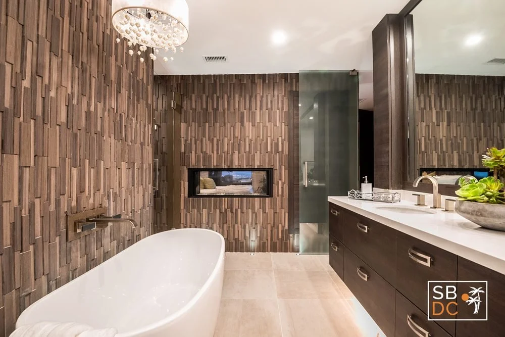 Modern bathroom with wood paneled wall, white freestanding bathtub, black vanity with white countertop, glass shower enclosure, and decorative plants.