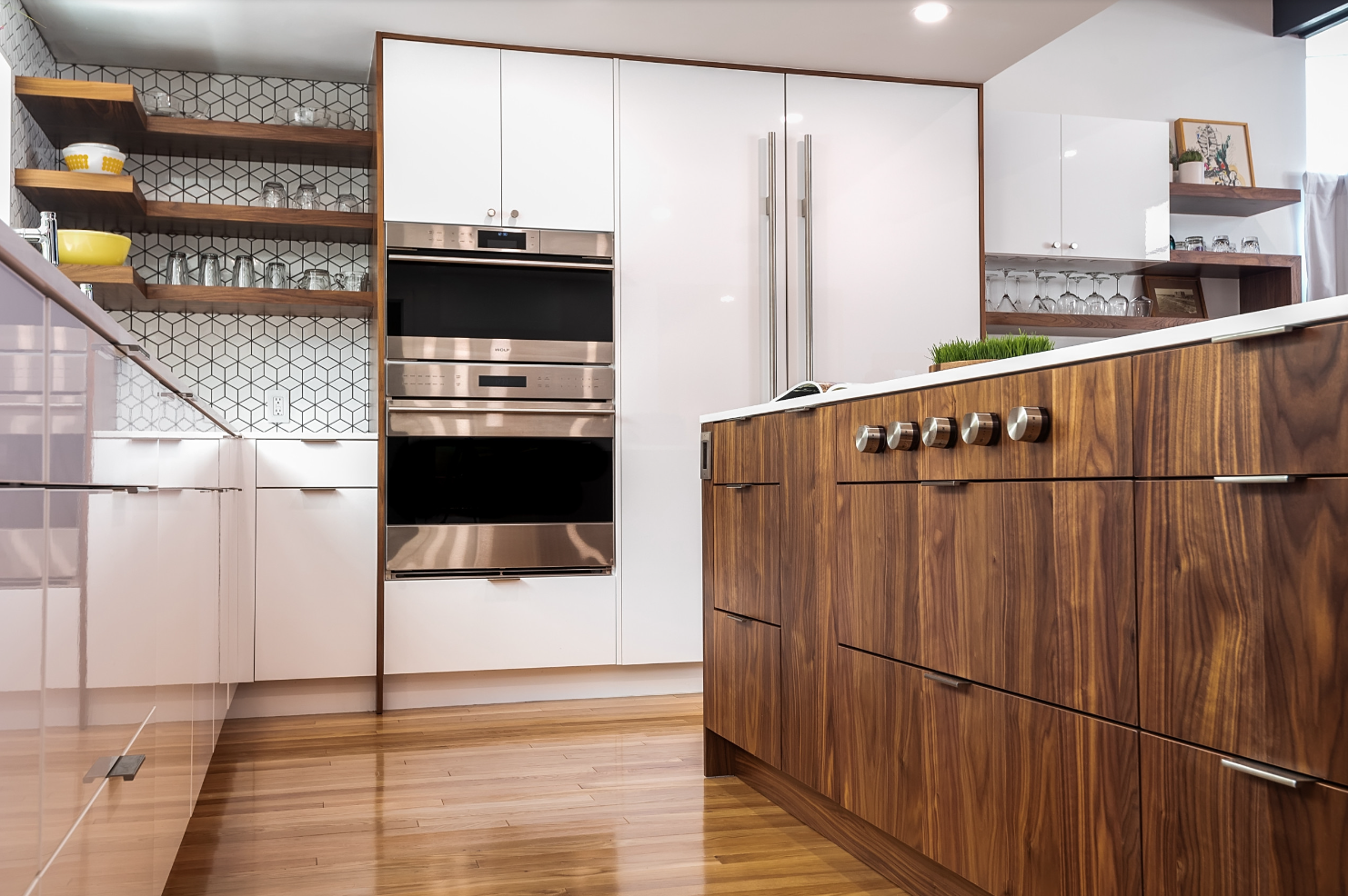 Modern kitchen with wooden and white cabinetry, open shelves with glassware, built-in stainless steel ovens, and a countertop with a small green plant.