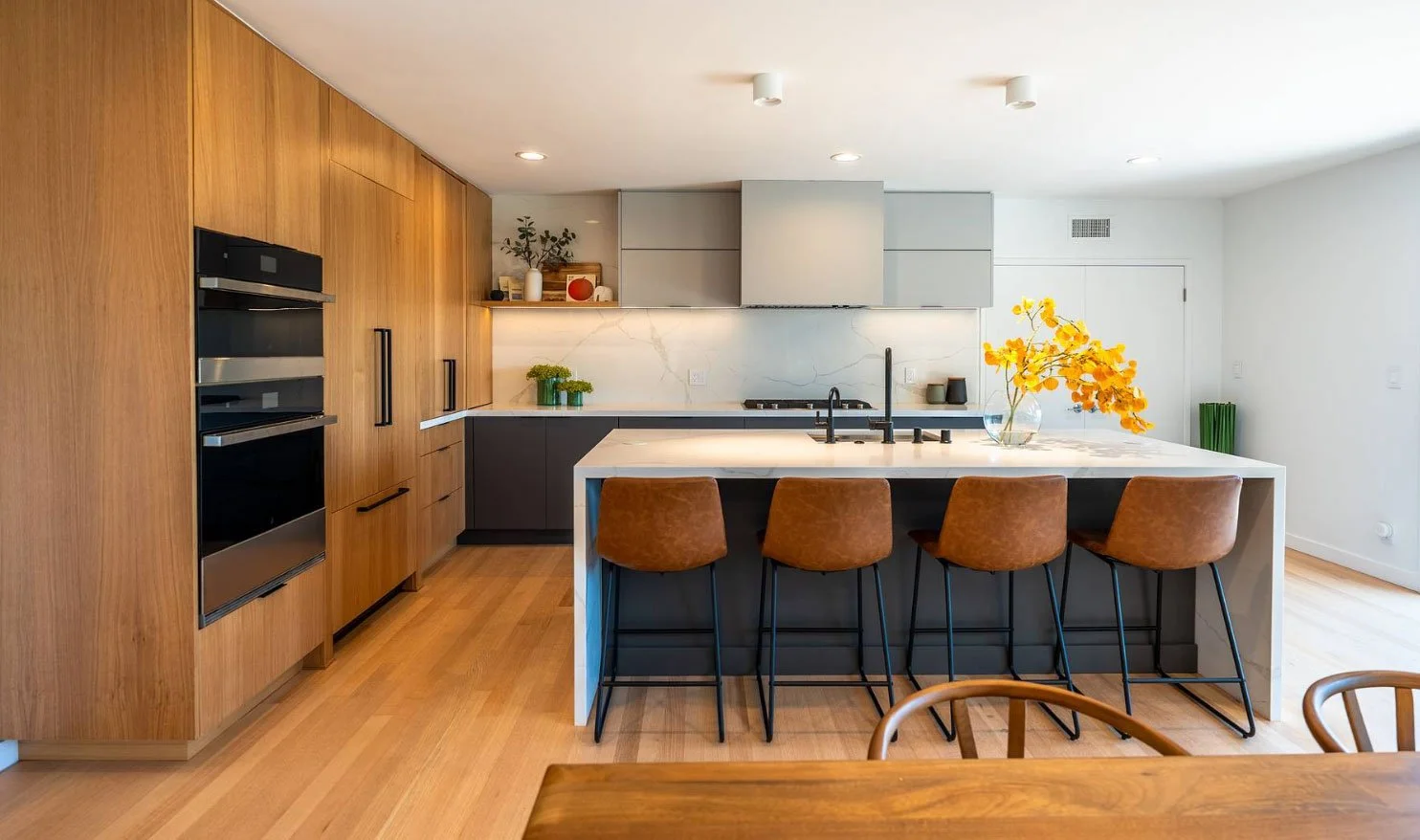 Modern kitchen with wooden cabinets, white marble backsplash, black and gray lower cabinets, kitchen island with brown chairs, and yellow flowers in a vase.