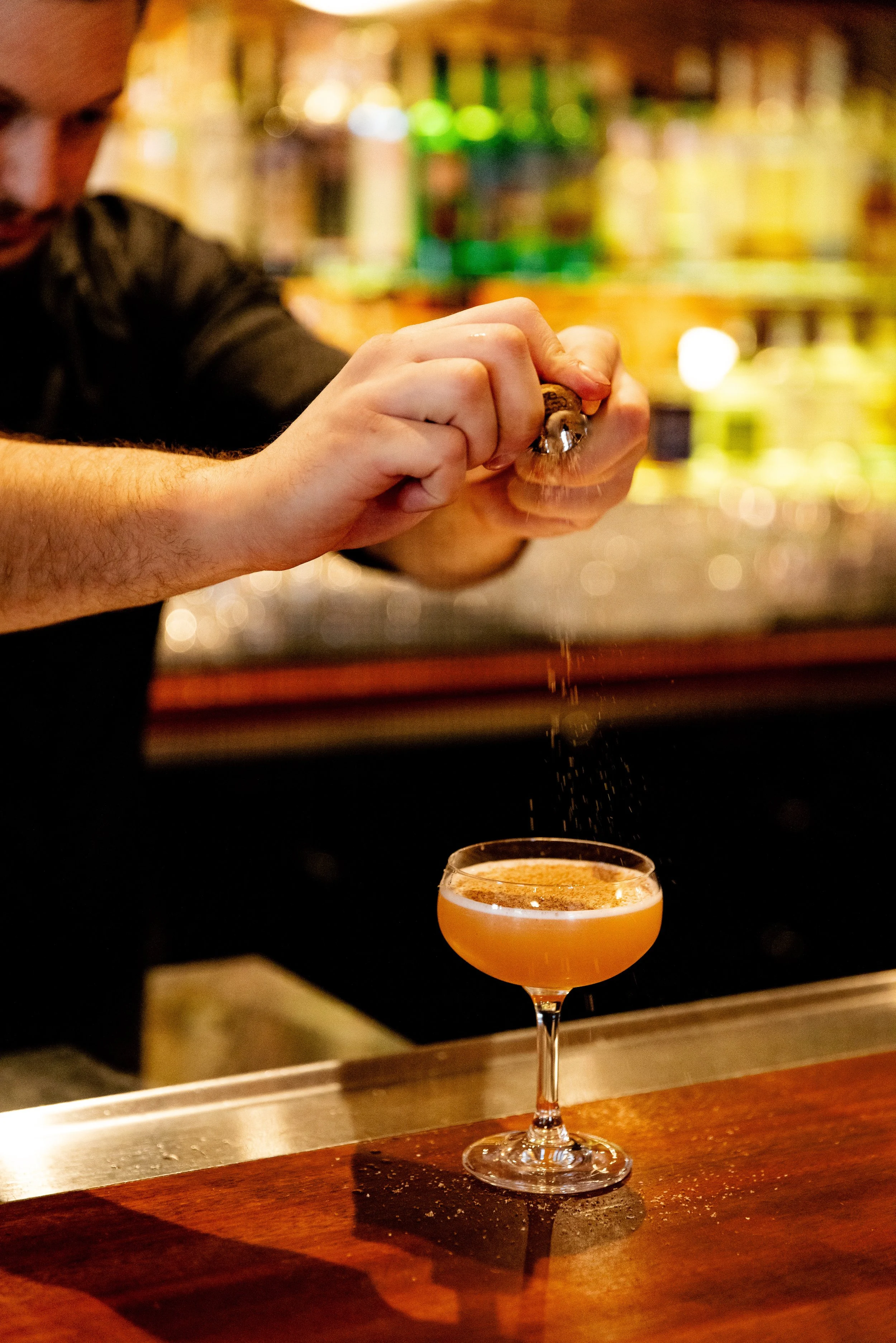 Barista sprinkles spice or garnish onto a cocktail in a coupe glass at a bar with colorful bottles in the background.