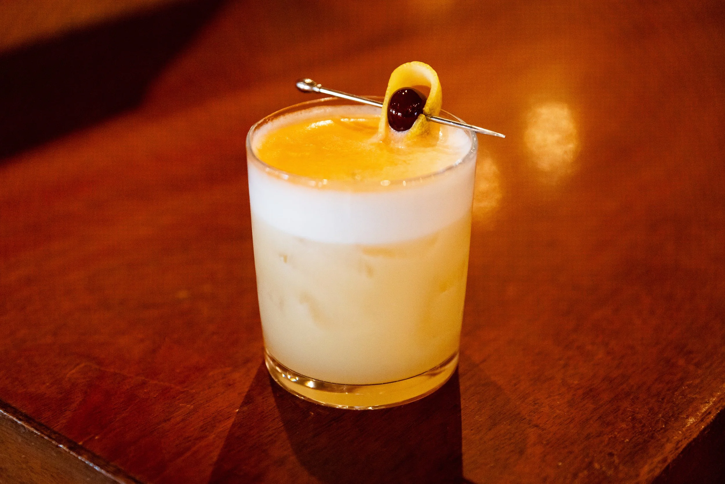 A cocktail with a creamy white layered appearance and a yellow-orange top, garnished with a lemon twist and a maraschino cherry, served in a clear glass on a wooden surface.