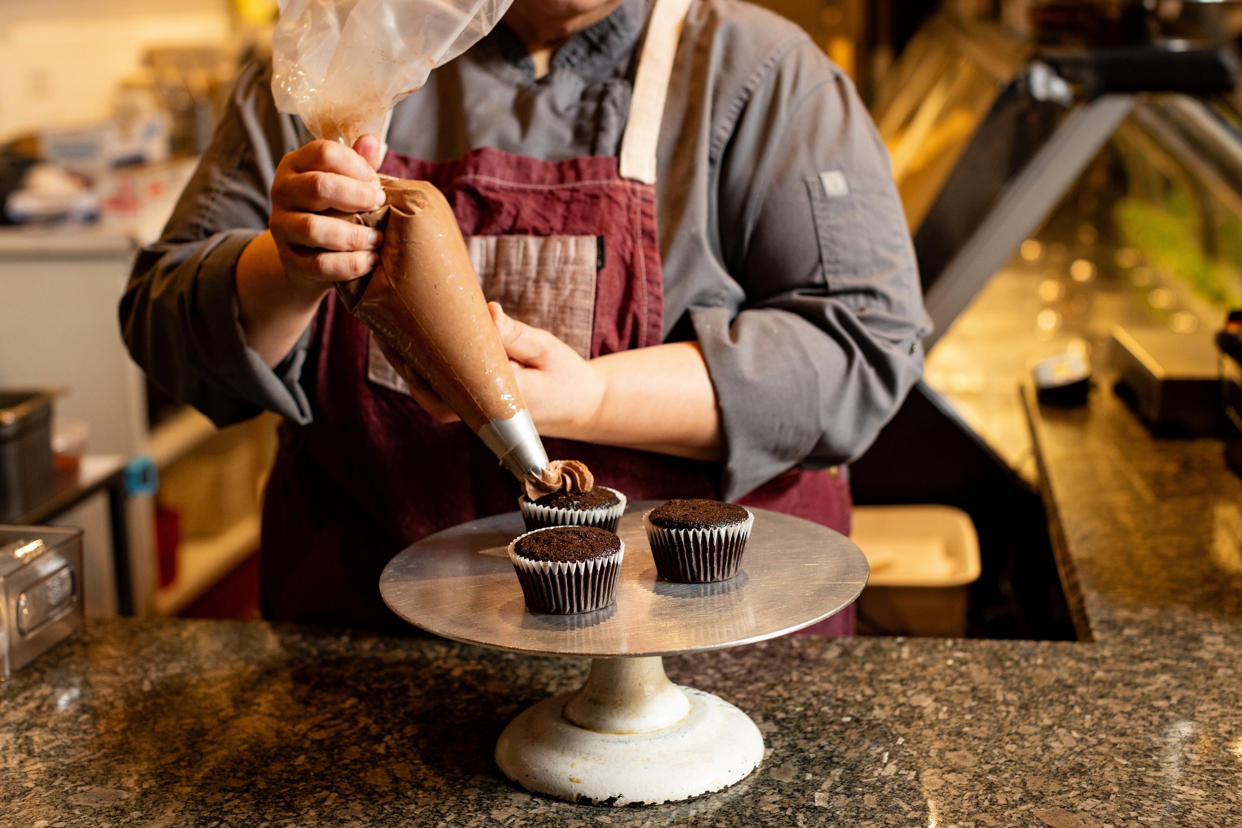 A person in a gray shirt and red apron is decorating cupcakes with chocolate frosting, using a piping bag, at a bakery or kitchen counter.