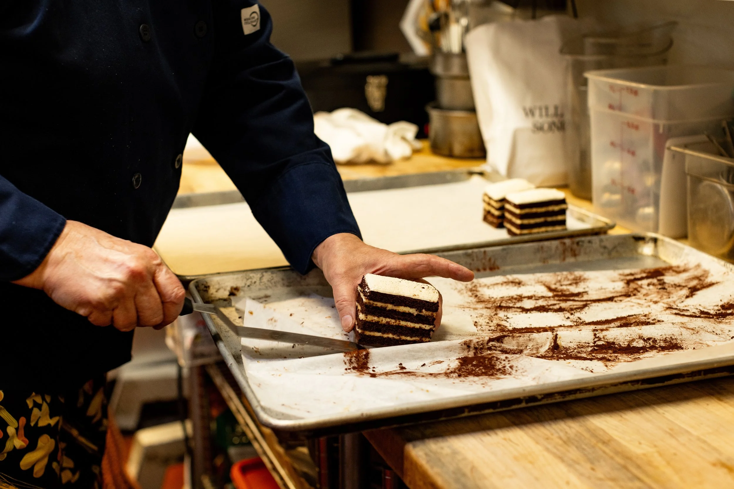 A person wearing a dark long-sleeve shirt is slicing a layered chocolate and cream dessert on a baking sheet dusted with cocoa powder, with a spatula.