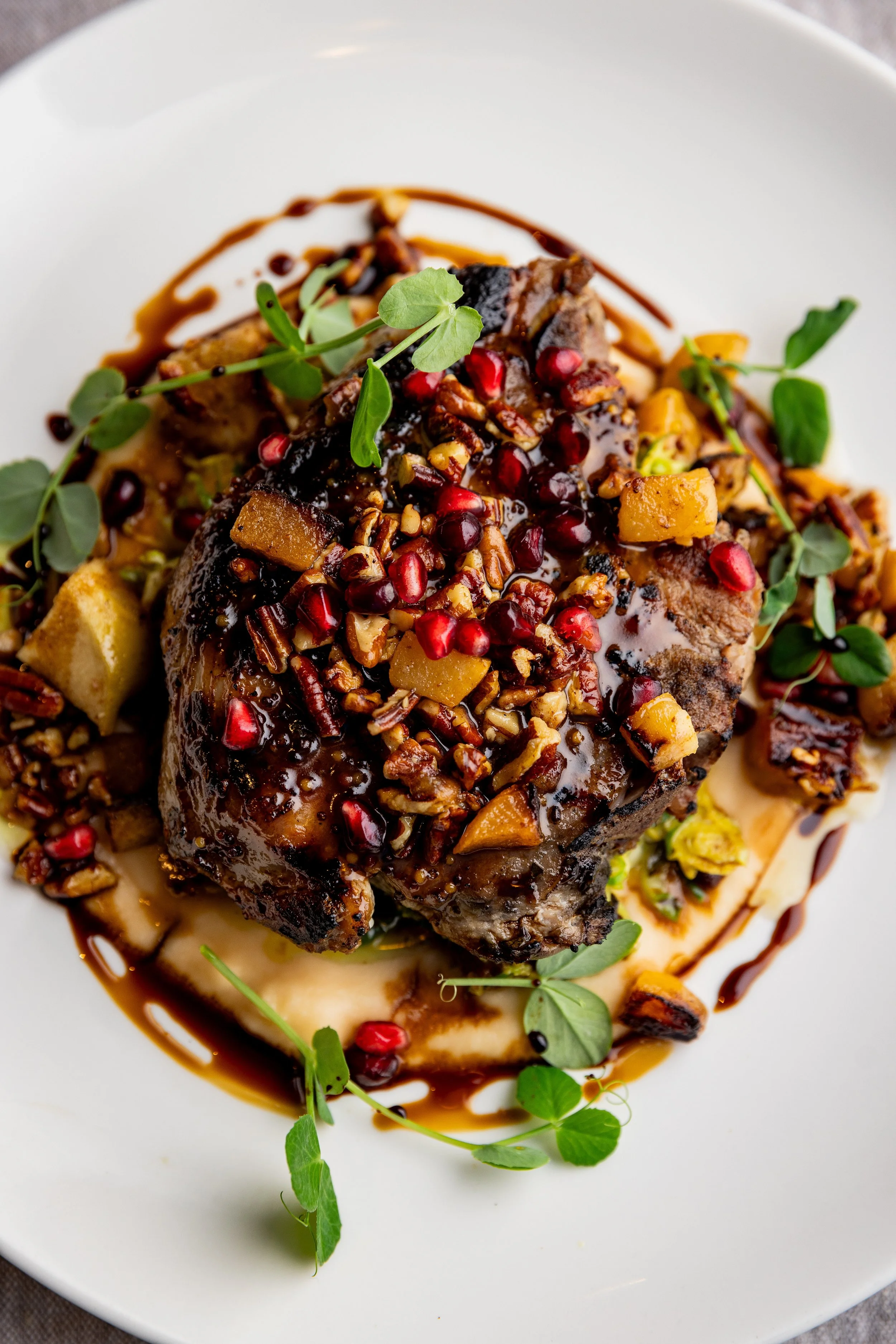 A plated dish featuring a cooked piece of meat topped with a sauce and garnished with pomegranate seeds, chopped nuts, diced vegetables, and fresh herbs, with drizzles of balsamic glaze and a puree underneath.