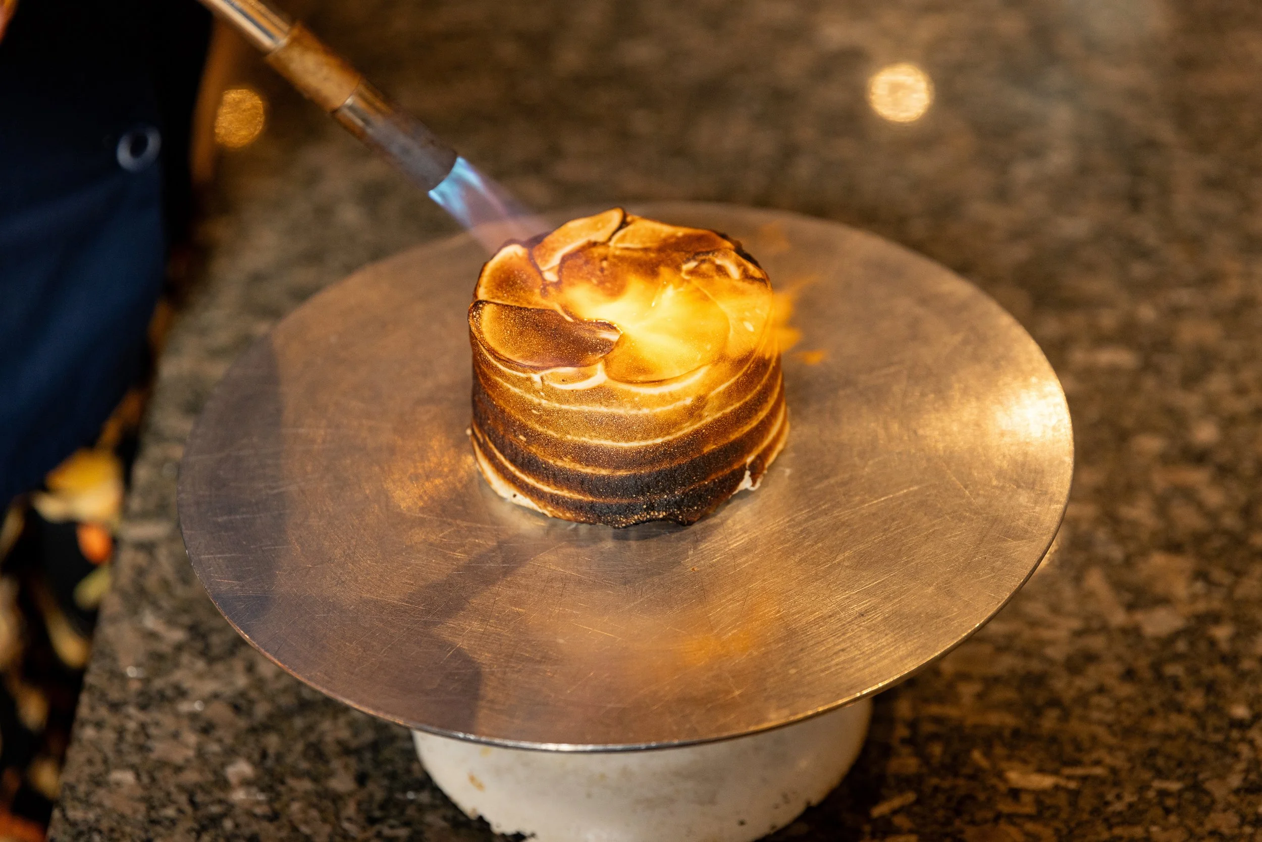 A dessert with torched marshmallow topping on a round, golden-brown cake, being caramelized with a blue-flame torch on a silver plate.